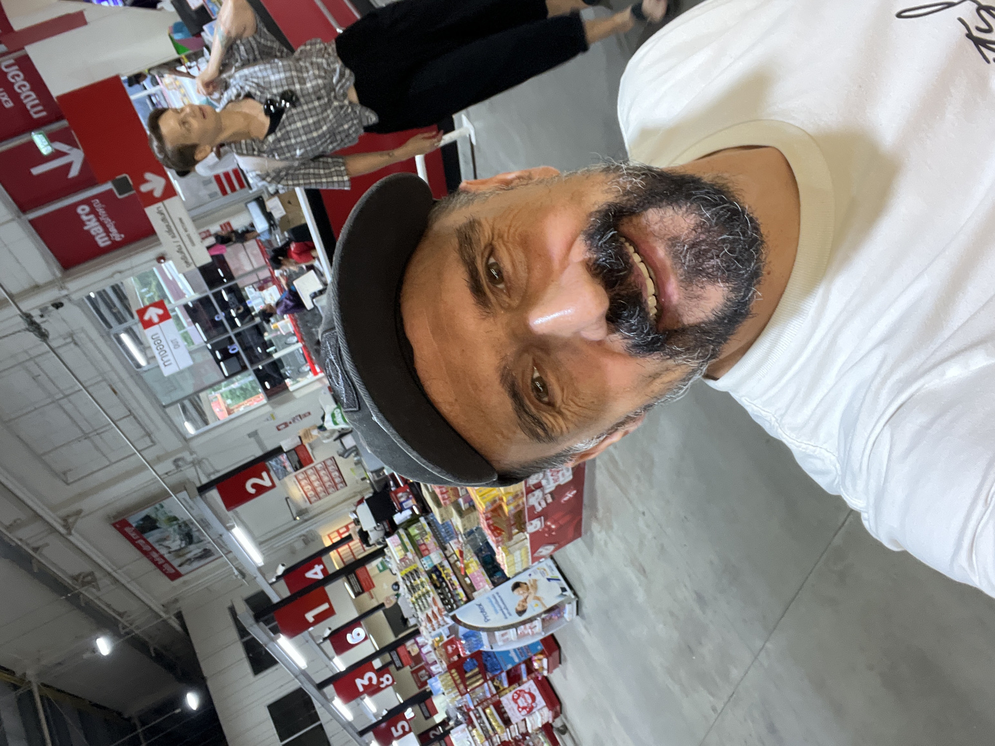 A man in a white shirt and a cap is taking a selfie in a Makro store. The aisles are marked with numbers 2, 5, 3, 8. Other signage with arrows points to exits. In the background, another individual in a plaid shirt and black pants is visible. The location is in Baan Bo Phut, Thailand.