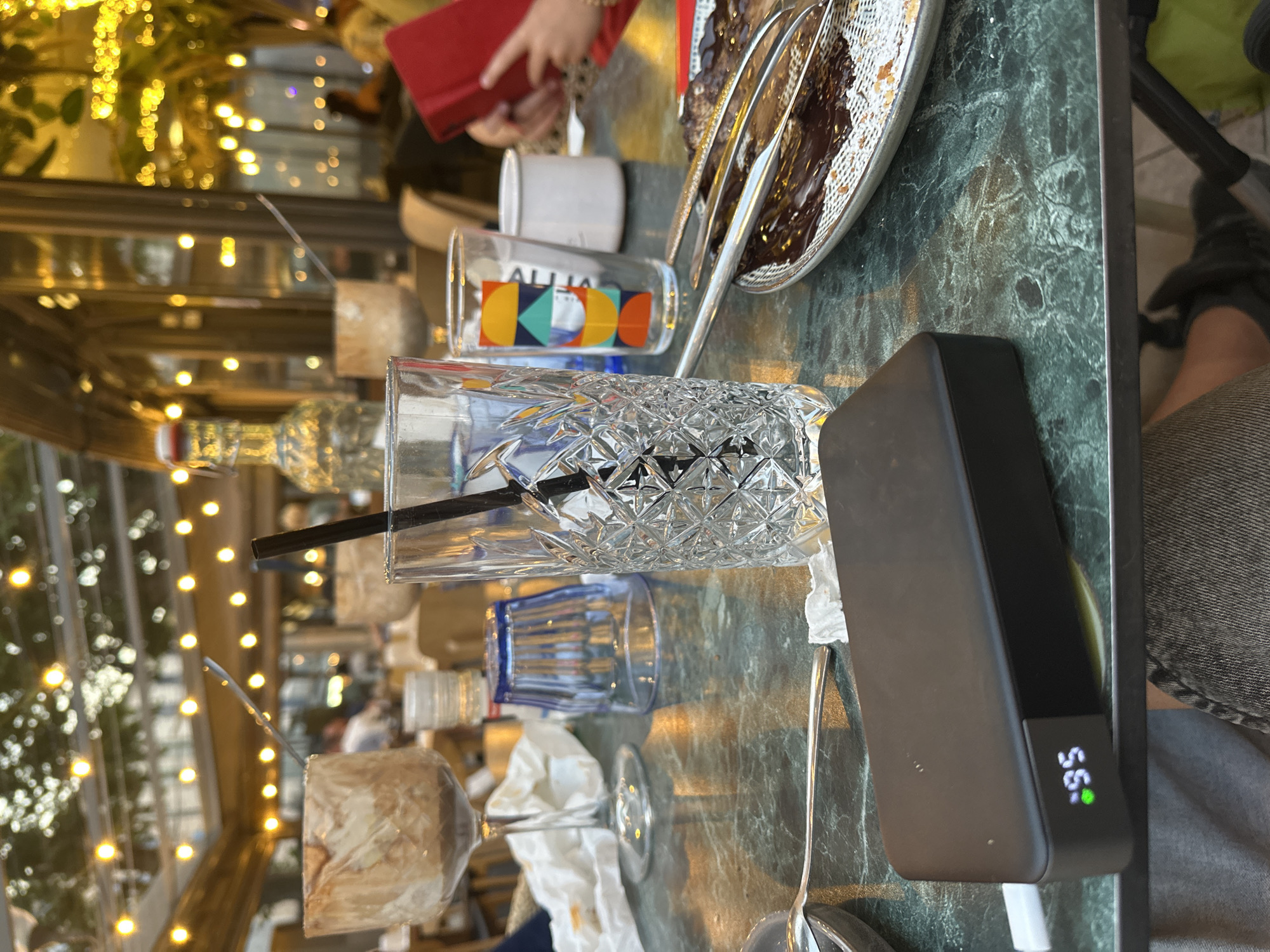 An outdoor table setting at a restaurant with a green marble tabletop is in focus. The table holds various items, including a glass with a black straw, a glass with colorful geometric shapes, a white cup, a red object that appears to be a book or phone case, and a plate with dessert. A black portable charger with a green digital display showing "95" is in the foreground. The background is blurred but shows more tables, lights, and people, suggesting a lively evening atmosphere. The overall scene appears to be in the city of Unknown, Unknown.