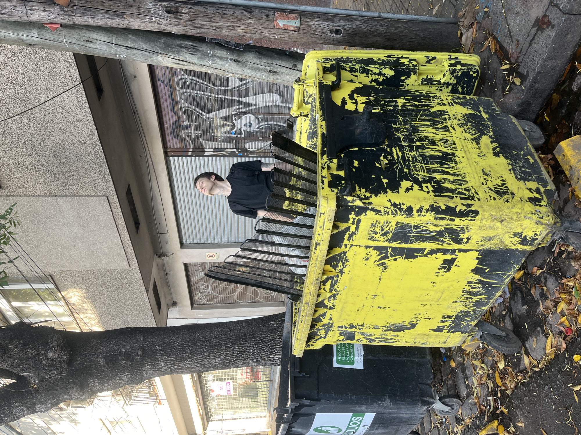 An urban street scene is depicted, dominated by a large, heavily worn yellow dumpster with black scratches and a black grate-like lid. A young man, wearing a black t-shirt and light pants, is positioned inside or behind this dumpster, with his eyes closed and head tilted upwards, conveying a sense of serenity or contemplation. His dark hair is short, and he has light stubble.
To the left of the yellow dumpster, a prominent, thick-barked tree trunk rises from the foreground. Below the yellow dumpster, a smaller black trash bin is visible, bearing a label with green accents. The ground is littered with fallen yellow and brown leaves, suggesting an autumnal setting.
In the background, various buildings line the street. To the left, a building features a textured, light-grey facade. Further back and to the right, another building displays a corrugated metal rolling shutter covered in graffiti, possibly indicating a closed shop. Overhead, two robust wooden utility poles extend vertically, with wires running between them. A small, faded red sign with a white arrow and the number "1" is attached to one of the poles.
Visible text on the black bin includes "RESIDUOS" (meaning "waste" in Spanish), followed by bullet points listing types of waste like "• RESIDUOS HU" (likely "HÚMEDOS" - organic waste), "• RESTOS DE CO" (likely "COMIDA" - food scraps), "• PAPELES SUCK" (likely "SUCIOS" - dirty papers), and "• CARTONES SU" (likely "SUCIOS" - dirty cardboard). The text "VIVAMOSVE" and "VICENTELOPEZO" (referencing Vicente López) are also visible, confirming the location in Olivos, Argentina. A red circular sign with an "E" on a building wall is also present. The scene is brightly lit, indicating daytime with clear weather.