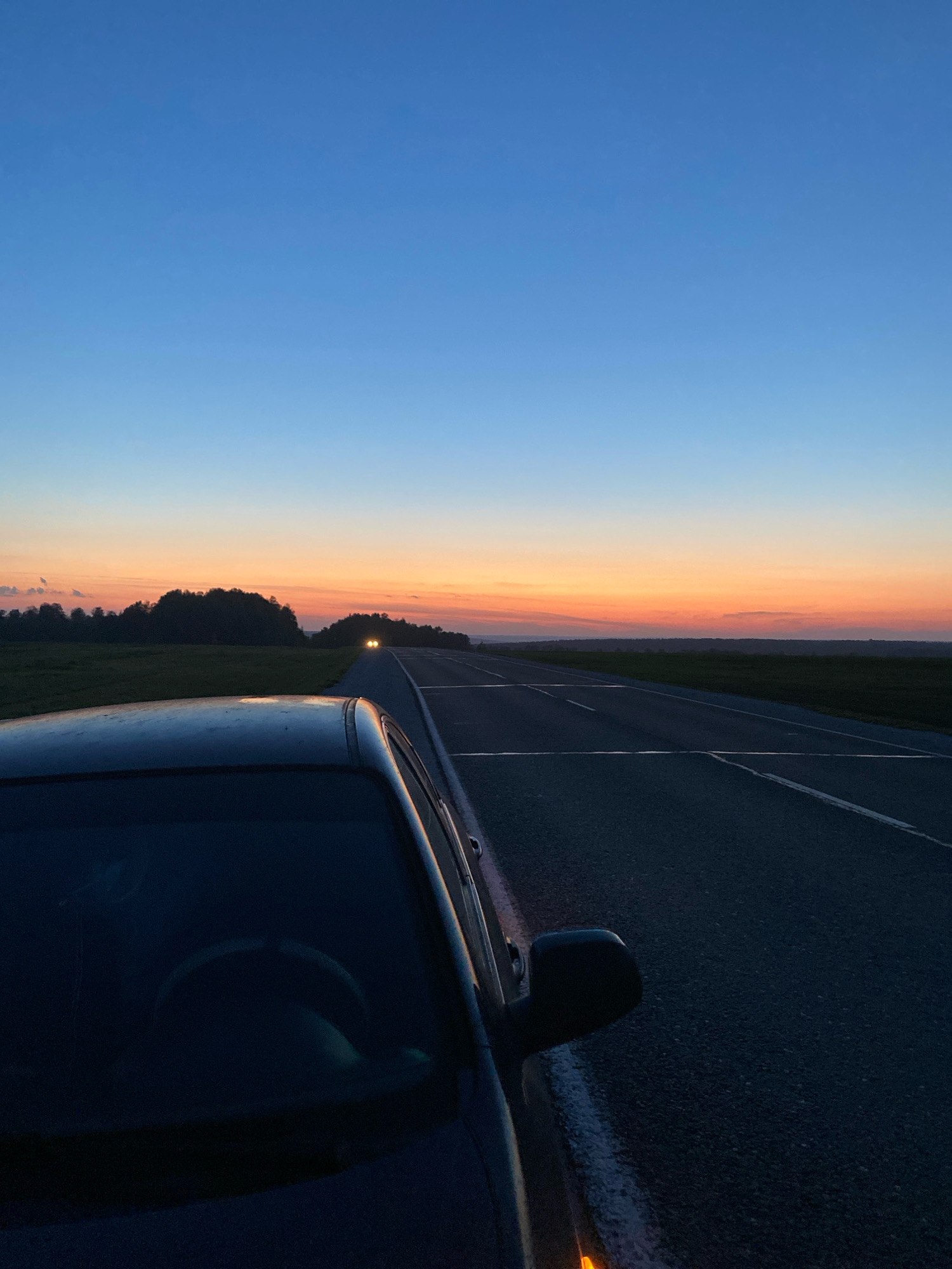 The image depicts a roadside scene at dusk, with a car parked on the right side of an asphalt road. The car's dark body and partially visible interior, including the steering wheel, are in the foreground. The road stretches into the distance, flanked by green fields and a treeline on the left horizon. The sky displays a gradient of colors, transitioning from deep blue at the top to orange and pink hues near the horizon, characteristic of a sunset. Two sets of headlights are visible in the distance on the road, indicating oncoming traffic. The setting appears to be a rural or semi-rural area, possibly on the outskirts of a settlement like Kolmogorovo, Russia, given the open landscape and the absence of urban infrastructure.