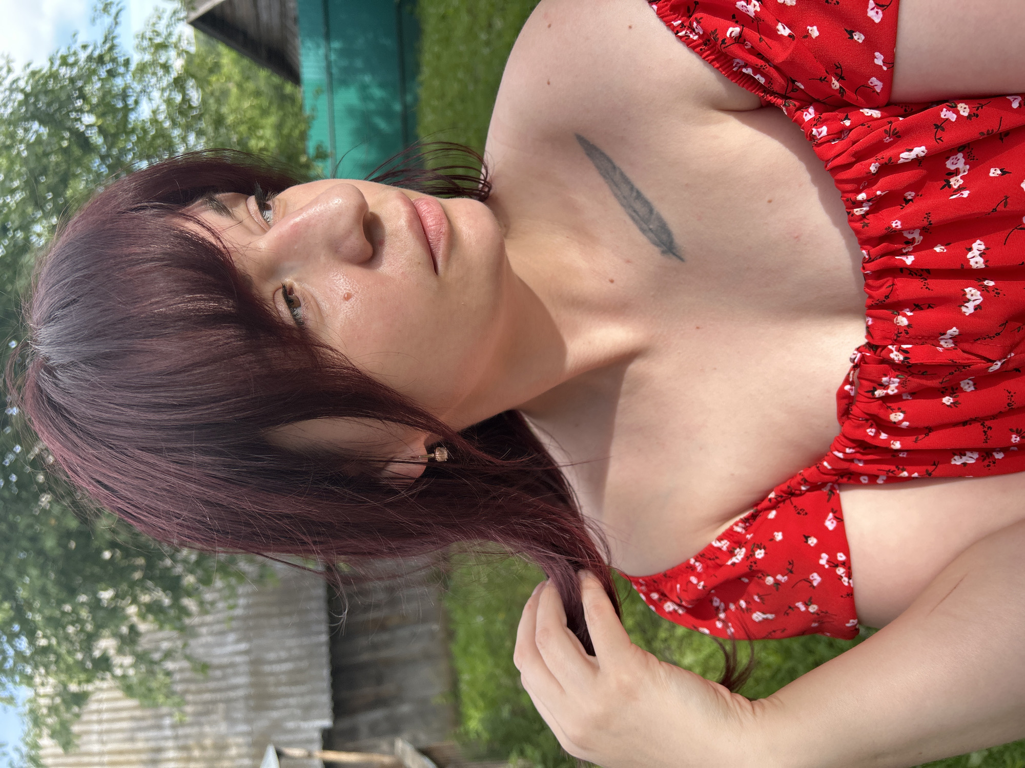 The image shows a medium close-up of a woman with dark red hair and bangs, wearing a red off-the-shoulder dress with a white floral pattern. She is outdoors, with a slightly out-of-focus background of green grass, trees, and rustic wooden structures, suggestive of a rural or suburban setting. The woman is looking to her right, with her left hand near her hair. A small feather tattoo is visible on her left shoulder. The lighting suggests it is daytime, possibly late afternoon, with sunlight casting shadows. The location is identified as Foky, Russia.