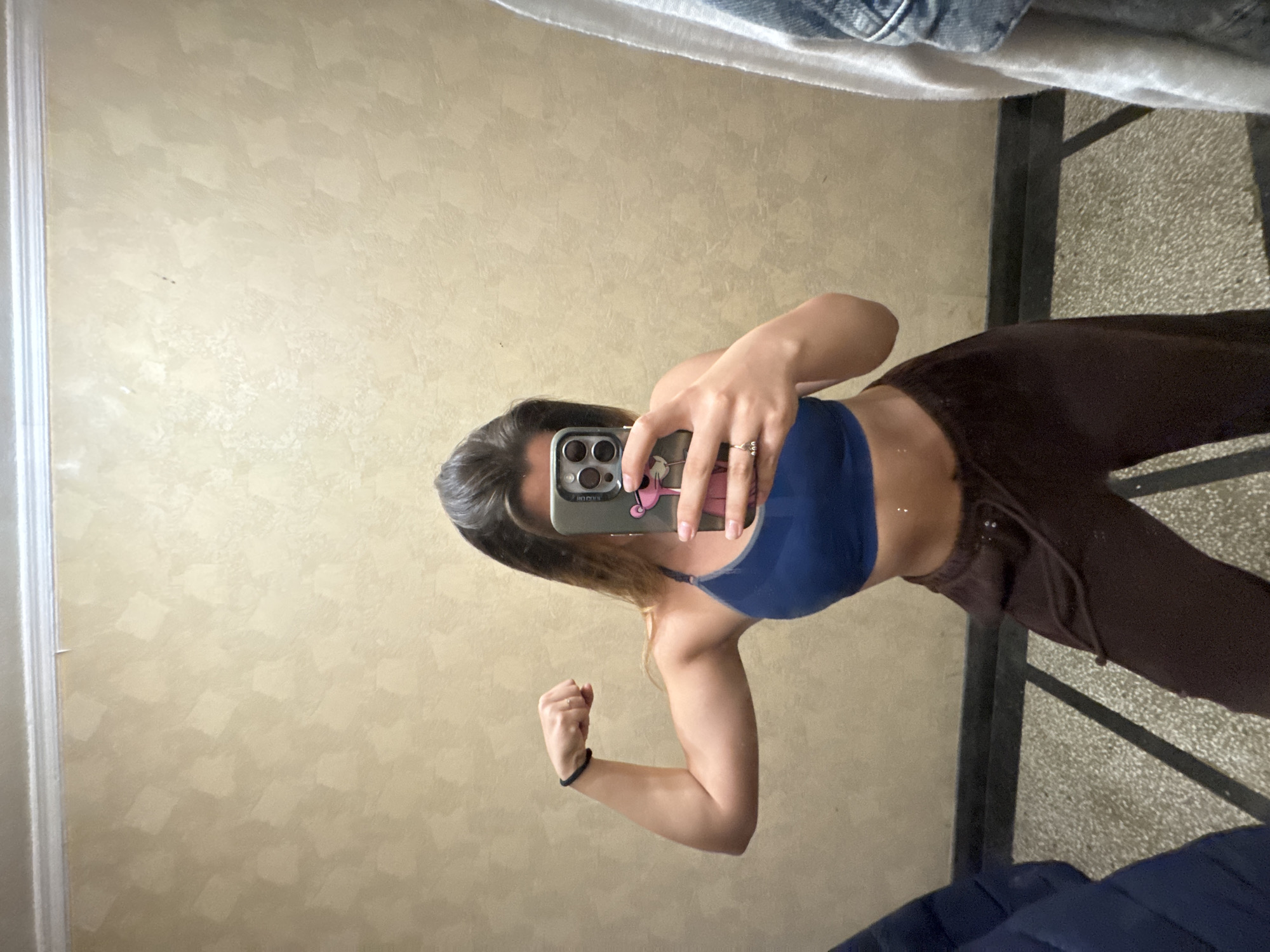 A woman is taking a selfie in a mirror. She is wearing a blue sports bra and brown sweatpants. She is flexing her left bicep. She has a black wristband on her left wrist. She is holding a cell phone with a patterned case. The background includes a beige wall, a silver metal rack, and a blue blanket. The location is not identifiable as Sofia, Bulgaria.