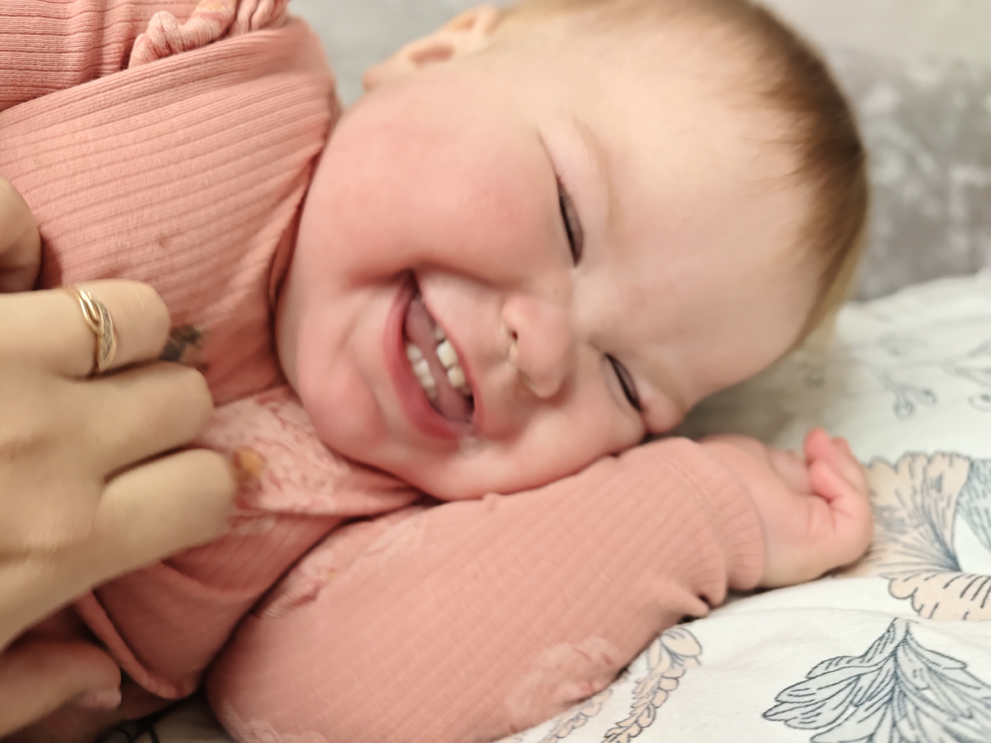This close-up, eye-level shot captures a joyful infant lying down, likely on a soft surface indoors. The infant has fair skin and light hair, and is wearing a ribbed, long-sleeved top in a salmon-pink or light-peach color. The child is caught mid-laugh or in a moment of intense happiness, with eyes squeezed shut, cheeks dimpled, and mouth wide open to reveal several tiny white teeth and gums. A small amount of drool or liquid is visible on the infant's chin.
An adult's hand is visible on the left side of the frame, gently resting on or touching the infant's arm or clothing. The adult wears a simple gold-toned ring with a decorative element on their ring finger. The background consists of a light-colored blanket or sheet adorned with a subtle floral or leafy pattern in muted grey-blue and light pink tones. The image conveys a strong sense of warmth, happiness, and affectionate interaction. No visible text is present in the image. The photograph was taken in Chaikovsky, Russia.