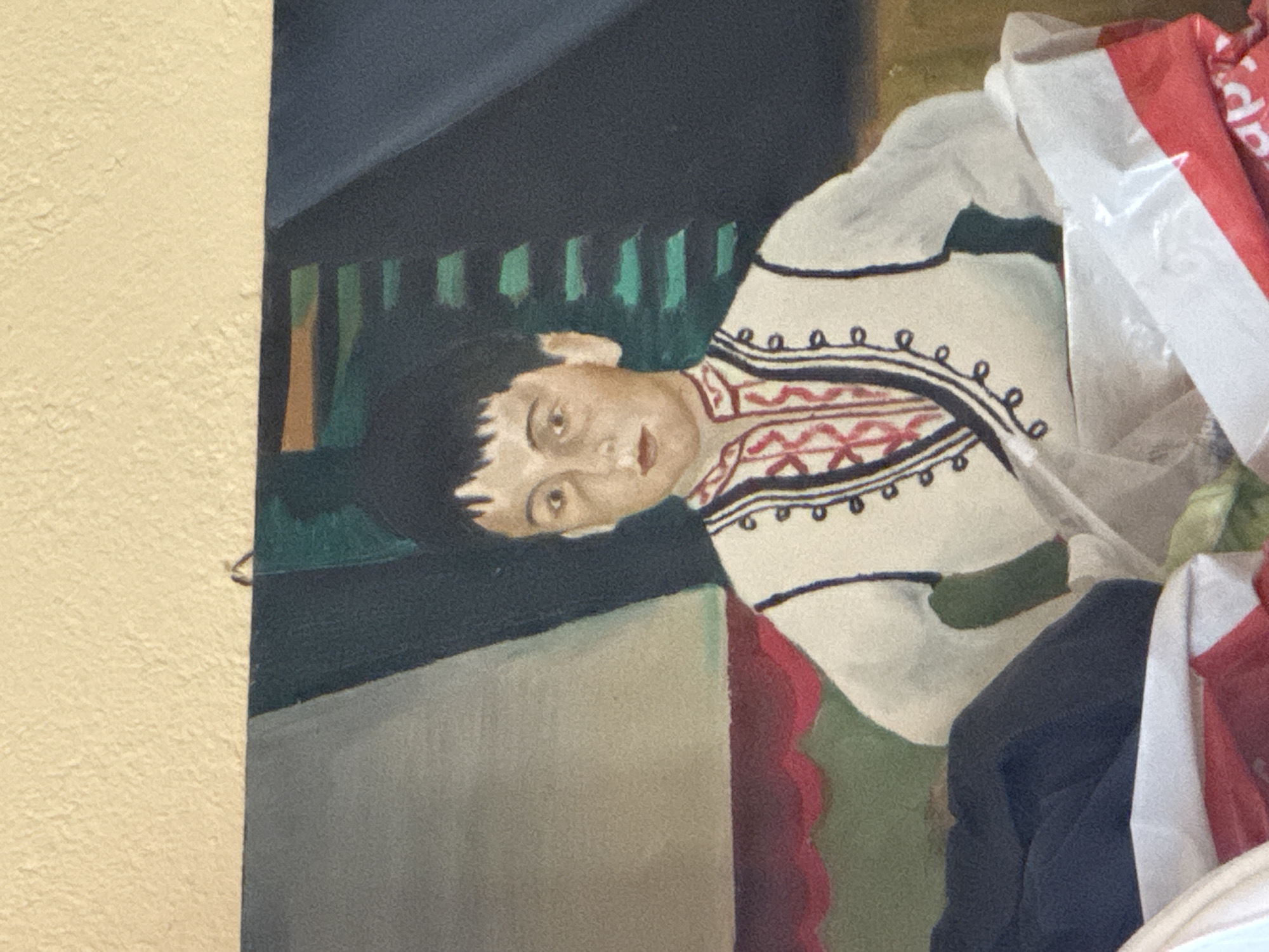 The image shows a painting of a young boy with dark hair, wearing traditional clothing with a white vest adorned with red and black embroidery. He has light skin and appears to be looking directly at the viewer. The background of the painting is abstract, with dark green and grey shapes. The painting is hanging on a textured, cream-colored wall. In the foreground, partially obscuring the bottom of the painting, are various items including a dark blue garment, and what appears to be a white plastic bag with red and white markings. The scene is indoors, with the painting being the primary subject. There is no indication of the city of Ruzhitsa, Bulgaria.