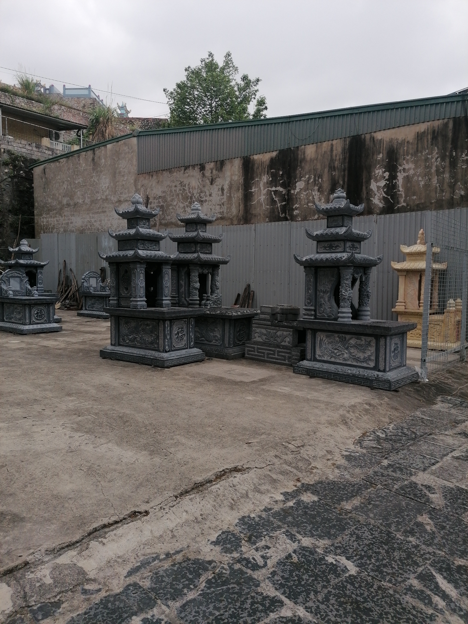 The image depicts a display of several traditional Asian tomb-like structures or memorials in what appears to be a sales or production yard. The structures are made of stone, primarily a dark grey, with intricate carvings. There are multiple pagodas and pedestals arranged on a weathered concrete surface. The setting seems to be outdoors, evidenced by the overcast sky and the presence of a building and wall in the background. The location is identified as Ha Tu Ward, Vietnam. The background consists of a weathered stone wall and corrugated metal fence, with some greenery visible above. There is a scattering of construction materials, pieces of wood and a wire cage.