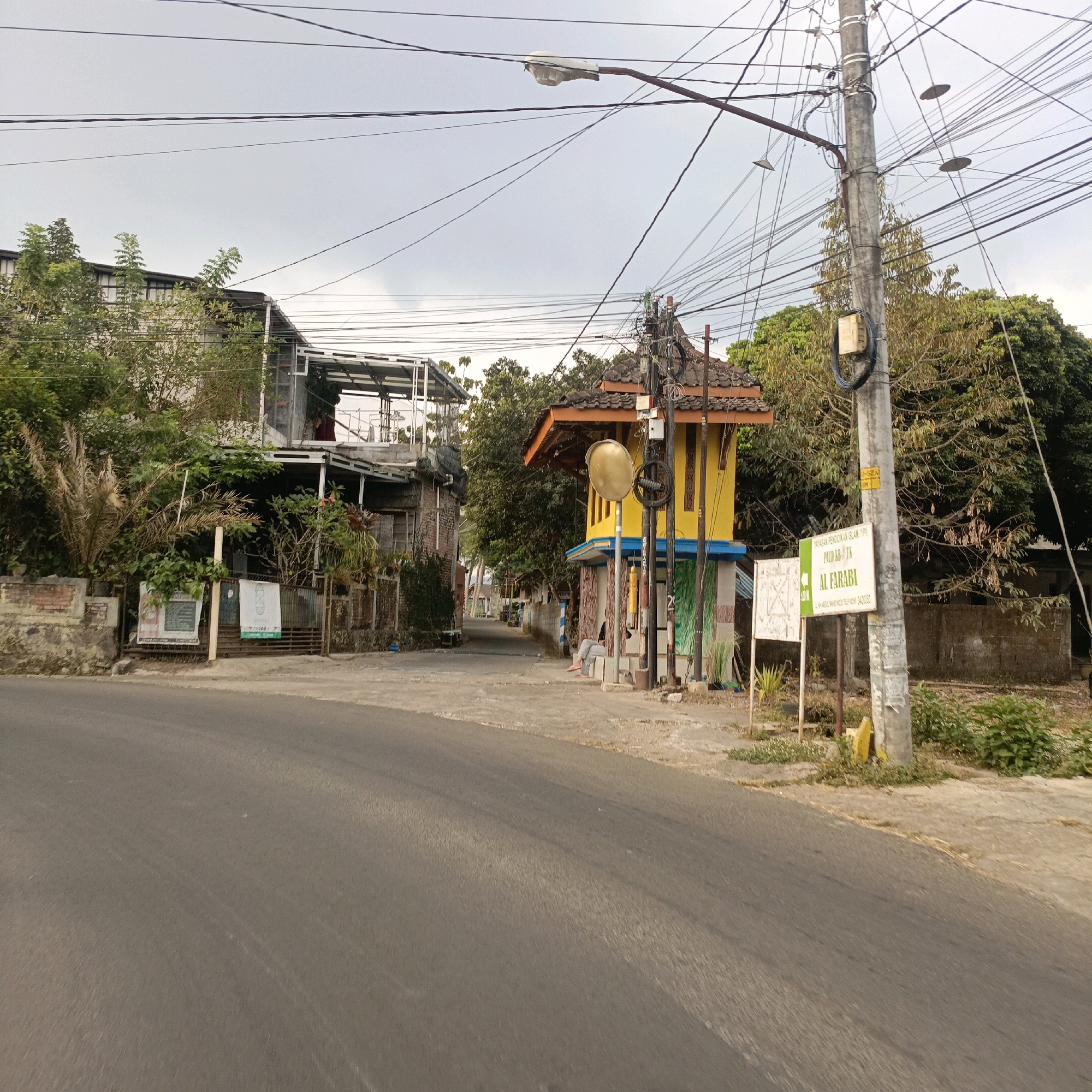The image captures a street scene in Salatiga, Indonesia, likely on a cloudy day. In the foreground, a paved road curves to the left. On the right side of the road, a tall utility pole is laden with numerous electrical wires, some of which are suspended over the street. Adjacent to the pole is a small, vibrant yellow structure with a brown tiled roof, possibly a kiosk or a small building. A convex mirror is mounted on a pole in front of this yellow structure. Further down the road, a two-story building with a metal frame and open sides is visible on the left, partially obscured by trees and foliage. The building on the left appears to be under construction or renovation. A sign with text is affixed to a fence in front of the building on the left. Behind the yellow structure, more trees and vegetation are present. The overall environment suggests a semi-urban or residential area with a mix of buildings and abundant greenery. A person is seated on a low wall or step in front of the yellow structure.