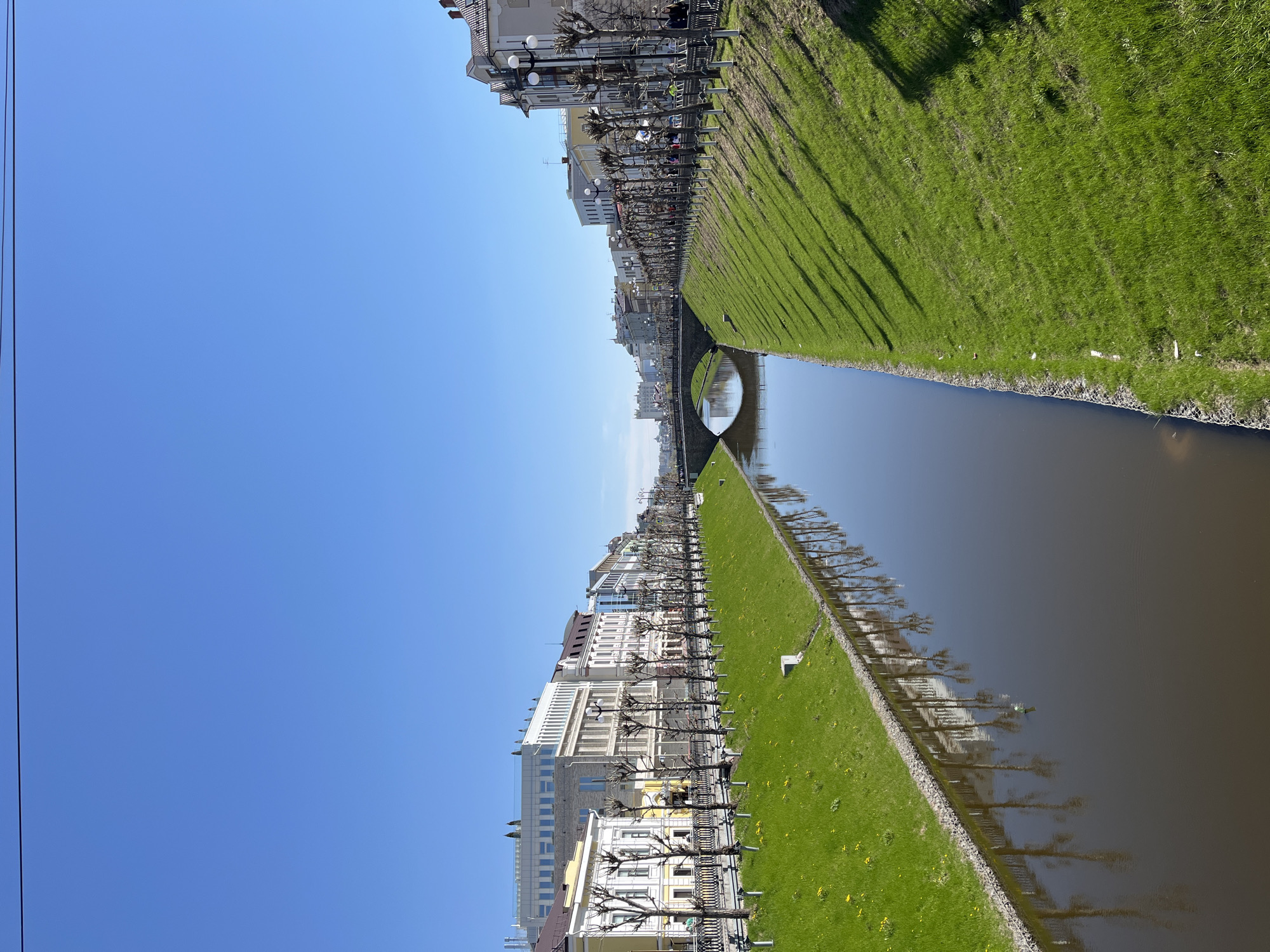 The image shows a canal in Kazan, Russia. Buildings line one side of the canal, interspersed with bare trees. A paved path runs along the buildings. On the other side of the canal is a green grassy bank. The canal water is dark, reflecting the buildings and trees. A small bridge crosses the canal in the distance. The sky is clear and blue. The scene is viewed from a high angle.
