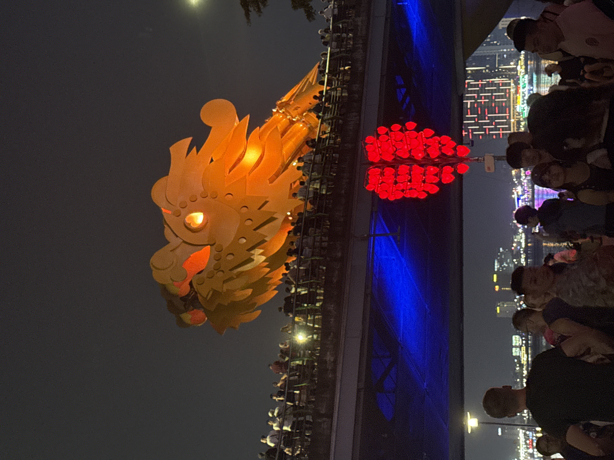 The image shows the Dragon Bridge in Đà Nẵng, Vietnam, at night. A large, illuminated dragon head is prominently displayed, seemingly attached to the bridge. Red light installations flank the dragon. People are visible on the bridge, observing the display. The city skyline, with illuminated buildings, is in the background. The environment appears to be a festive or celebratory event at night. Blue lights also appear to illuminate sections of the bridge.