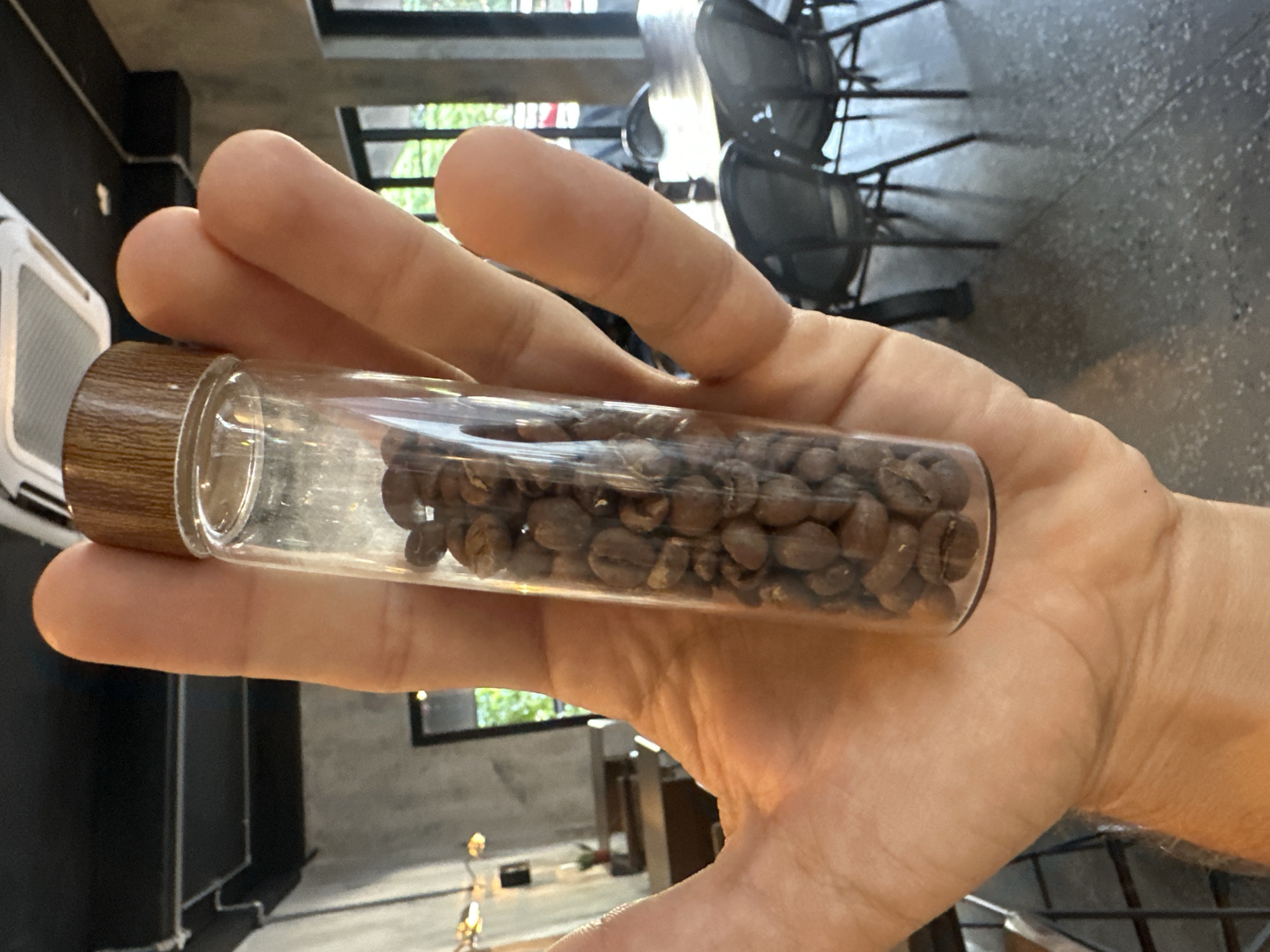 A close-up, waist-up, interior shot shows a person's left hand holding a clear glass vial filled with roasted coffee beans. The vial has a wooden cap and is positioned in the center of the frame. The background reveals a dimly lit coffee shop with concrete walls, black ceiling beams, and tables with chairs. A bright light fixture is visible on the ceiling, and natural light filters in from windows at the rear. The overall ambiance suggests a modern cafe environment in Đà Nẵng, Vietnam.