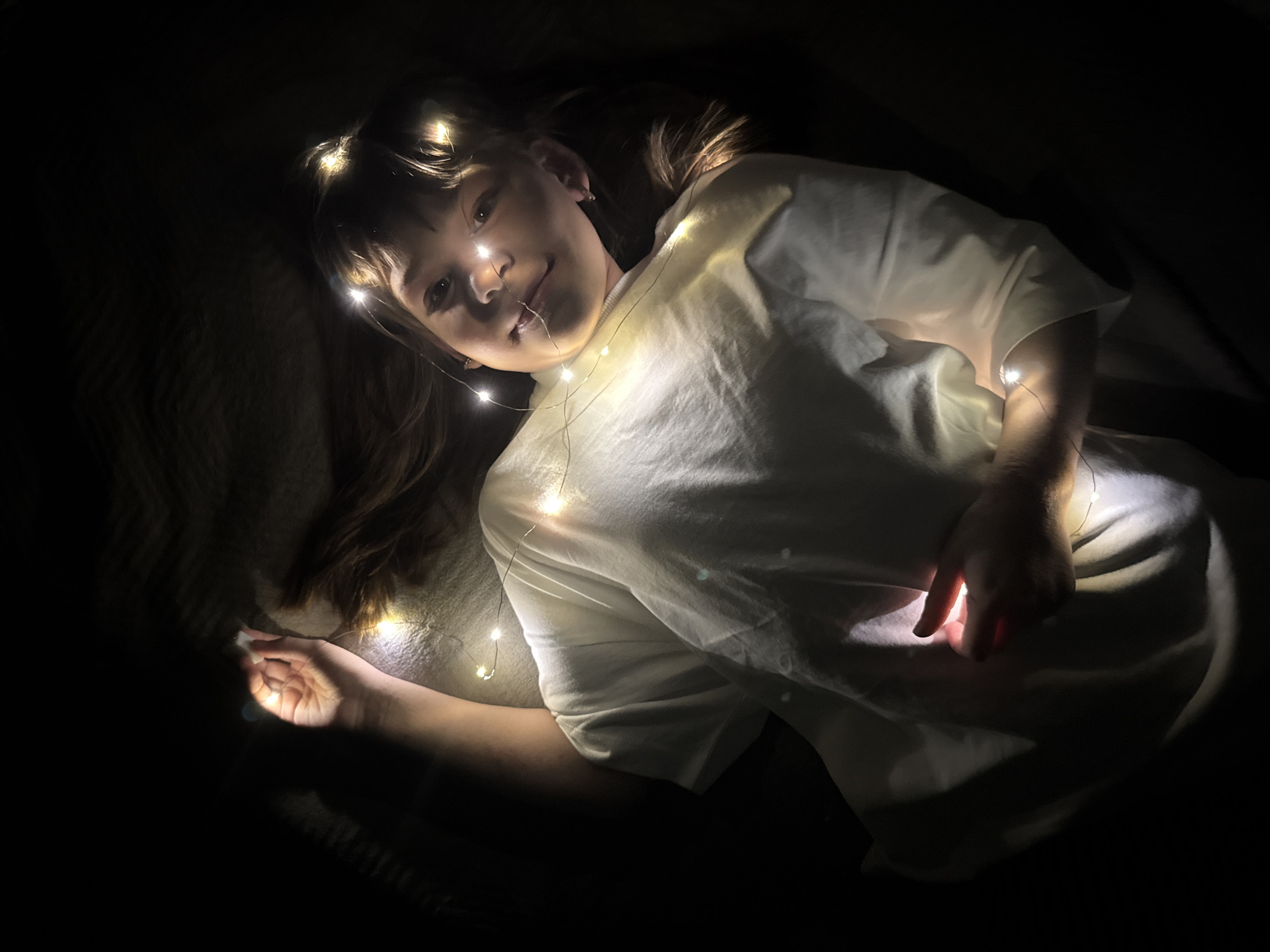A girl is lying down in a dark room, covered with a string of fairy lights. Her head is resting on a soft surface, possibly a pillow or a blanket. She is wearing a white t-shirt. The string lights are draped across her body and around her face, illuminating her features. The background is dark and indistinct. The location in the city of Rusanovo, Russia cannot be determined from this image.