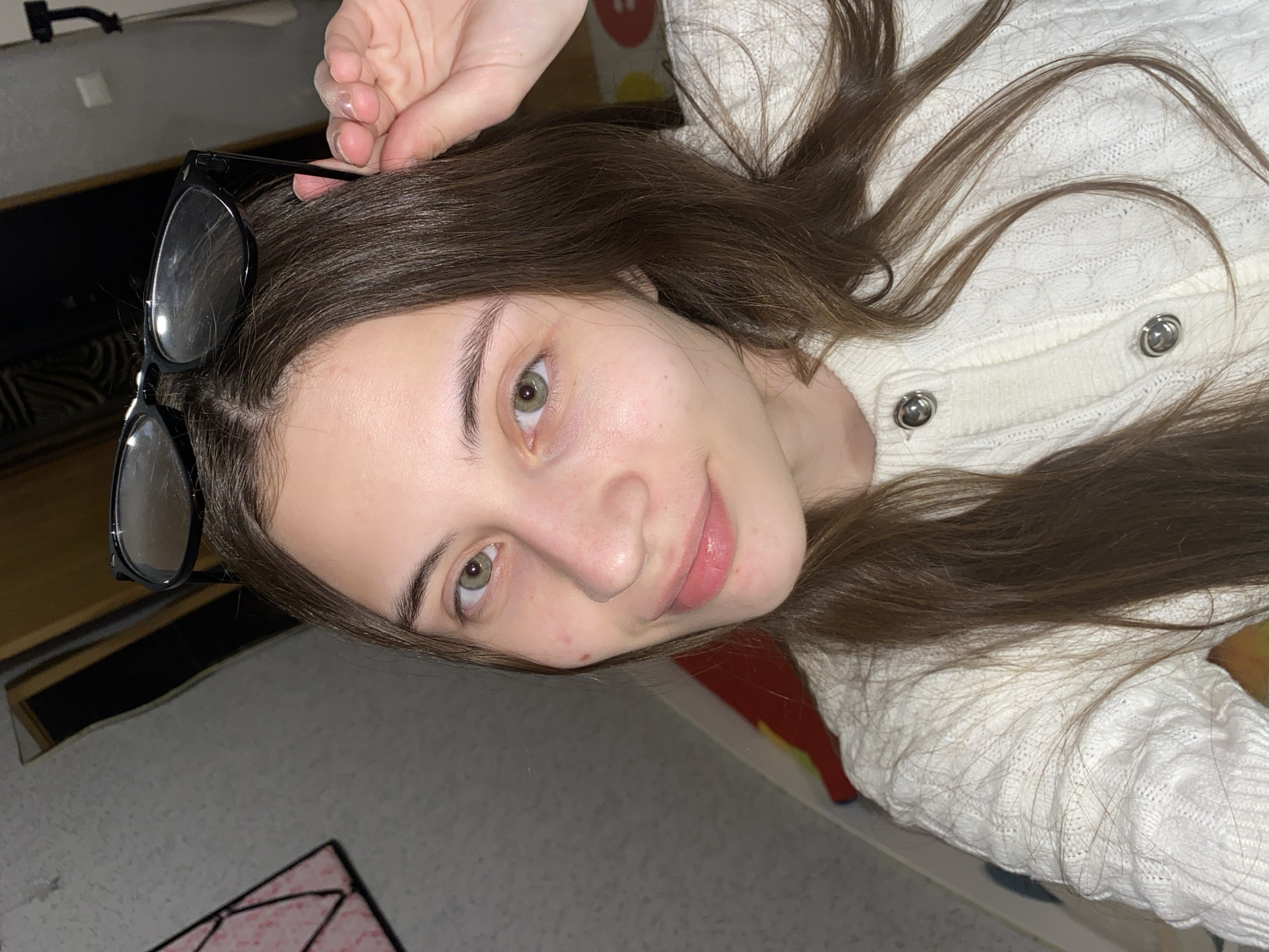 A young woman with long, dark brown hair and green eyes is depicted in a close-up, indoor portrait. She appears to be lying down, with her hair spread out around her head and shoulders. Her light complexion shows a few minor blemishes, and she has a gentle, soft smile on her lips. She is wearing a white, cable-knit cardigan with round silver buttons. A pair of black-framed glasses rests on top of her head, nestled in her hair, with her right hand visible near the frames, seemingly adjusting or holding her hair.
The background suggests an indoor setting, with a light-colored, possibly carpeted floor or rug visible. To the left, a dark, indistinct object, possibly furniture, is in the background, along with a small portion of a pink and white patterned mat. On the right, a colorful, possibly red and yellow, object is partially visible. The lighting is artificial, indicating an interior space. The woman's expression is calm and pleasant. There is no visible text in the image. The location context provided is Votkinsk, Russia.