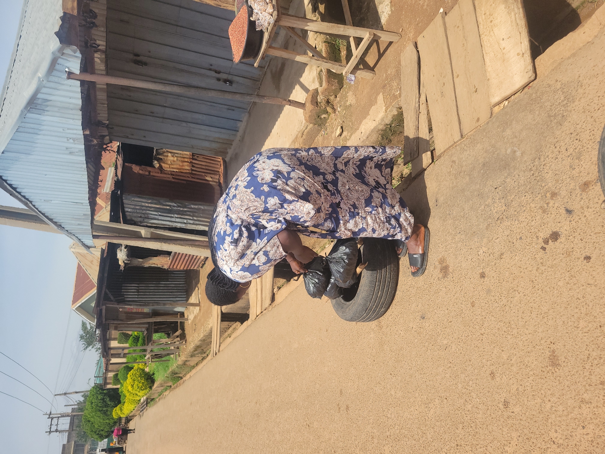 In Jos, Nigeria, a woman in a blue and white floral dress is seen standing on a paved road next to a ditch with wooden planks crossing it. She is bent over, tending to several black plastic bags of what appears to be trash placed inside an old tire. The scene is lined with small stalls made of corrugated metal and wood, with some displaying goods like grains and bagged items. Vegetation, including manicured green bushes and some trees, is visible along the roadside. The weather appears to be sunny and clear.