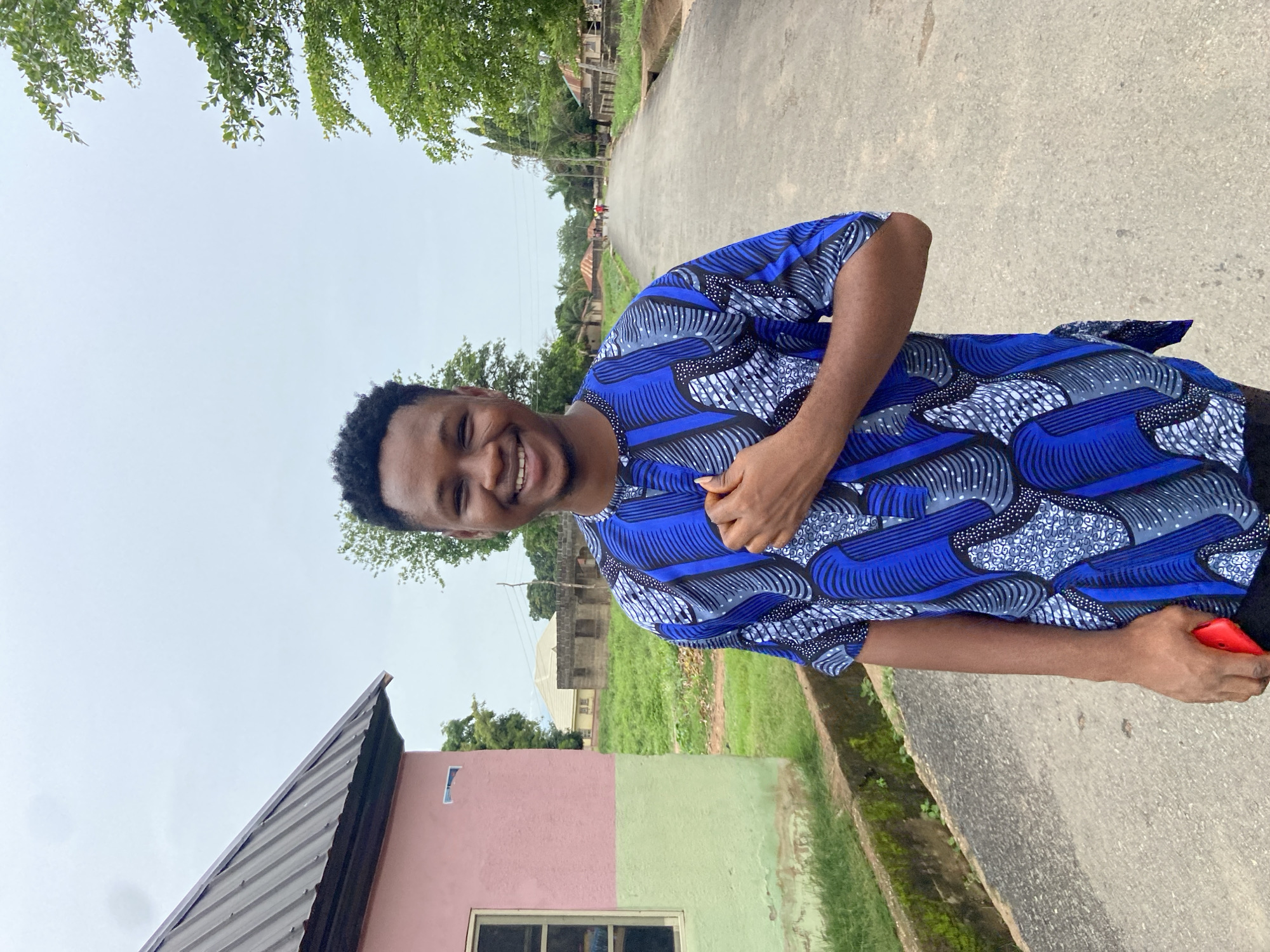 A young man is standing outdoors on a gray concrete road or pathway, smiling and looking at the camera. He is wearing a short-sleeved shirt with a blue, white, and gray patterned design. In his right hand, he appears to be holding a red object, possibly a phone.
Behind him is a scene typical of Mararaba, Nigeria, featuring a mix of residential and natural elements. There are buildings of various designs and construction styles visible, some with roofs and walls in shades of pink and green. The houses appear to be spread out, with patches of green grass and trees dotting the landscape. The sky is overcast, suggesting a cloudy day. In the distance, there are more buildings and trees lining the road, indicating a residential area or small village.
