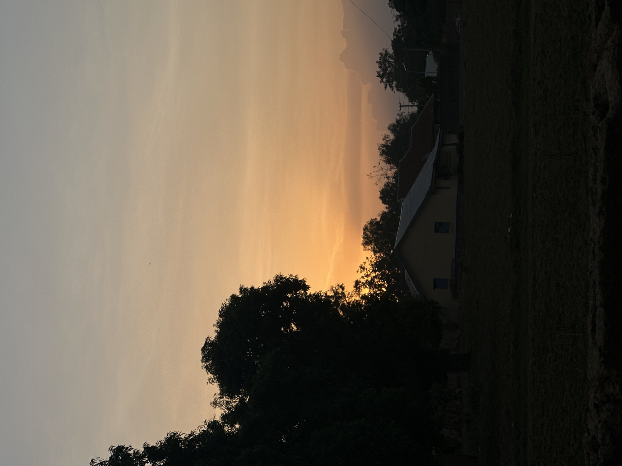The image shows a landscape in Bukuru, Nigeria, at dusk or dawn. The sky is a gradient of pale gray to warm orange. A large tree dominates the foreground, obscuring the lower portion of the horizon. A building with a slanted roof is visible behind the tree, likely a house. The ground appears to be covered in grass or vegetation. The overall impression is one of a rural or semi-rural setting at twilight. There are no people visible in the image.