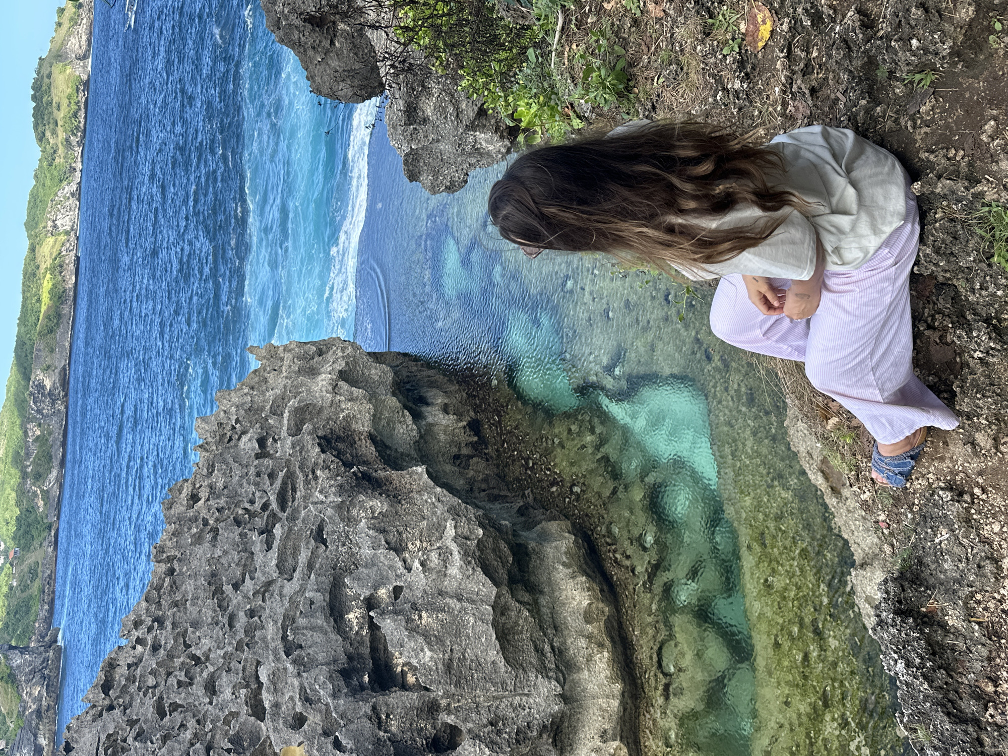 The photograph depicts a young person seated on a rocky cliff overlooking a body of water in Nusa Penida, Indonesia. The person, with long brown hair, is wearing a light-colored shirt and striped pants, and sandals. They are seated near the edge of the cliff, looking down at the turquoise water. The cliff is made of porous gray rock. Beyond the calm water, there is a section of open ocean with blue waves. Vegetation is visible along the cliff edge. In the background, a coastline can be seen under a clear sky.