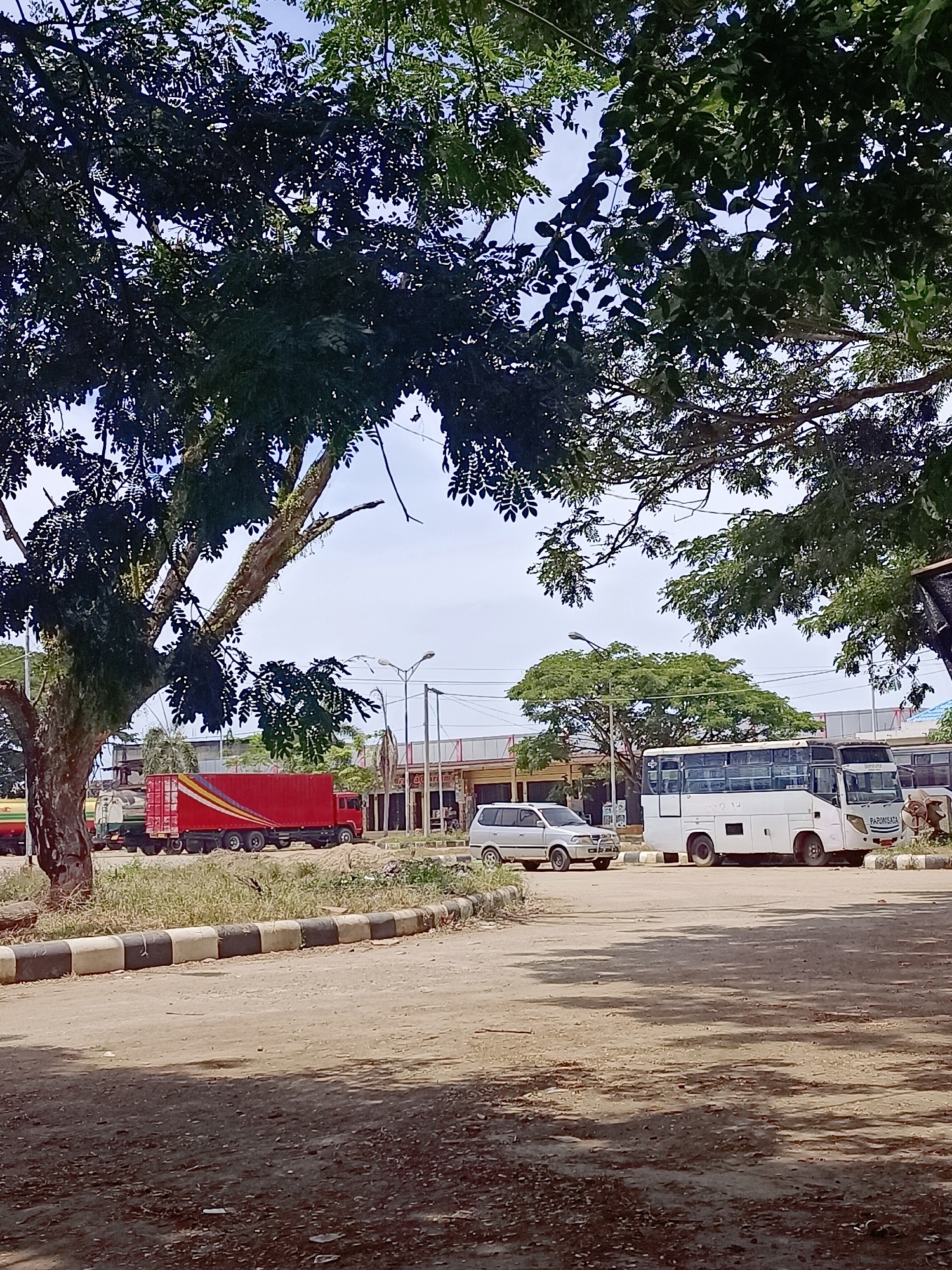 The image shows an outdoor scene with vehicles parked on a dirt and gravel lot under a canopy of trees. In the foreground, a dirt road with a black and white striped curb is visible. In the background, several vehicles are parked, including a red shipping container truck with yellow and red stripes, a white SUV, and a white bus with "PARIWISATA" written on it. The bus appears to be a passenger bus. Further in the background, there are buildings and other vehicles that are less distinct. The sky is overcast. The scene is likely a bus terminal or a truck depot in Meunasah Pupu, Indonesia, given the context. There are no people visible in the image. The environment appears to be a semi-urban or rural area with vegetation and trees providing shade.