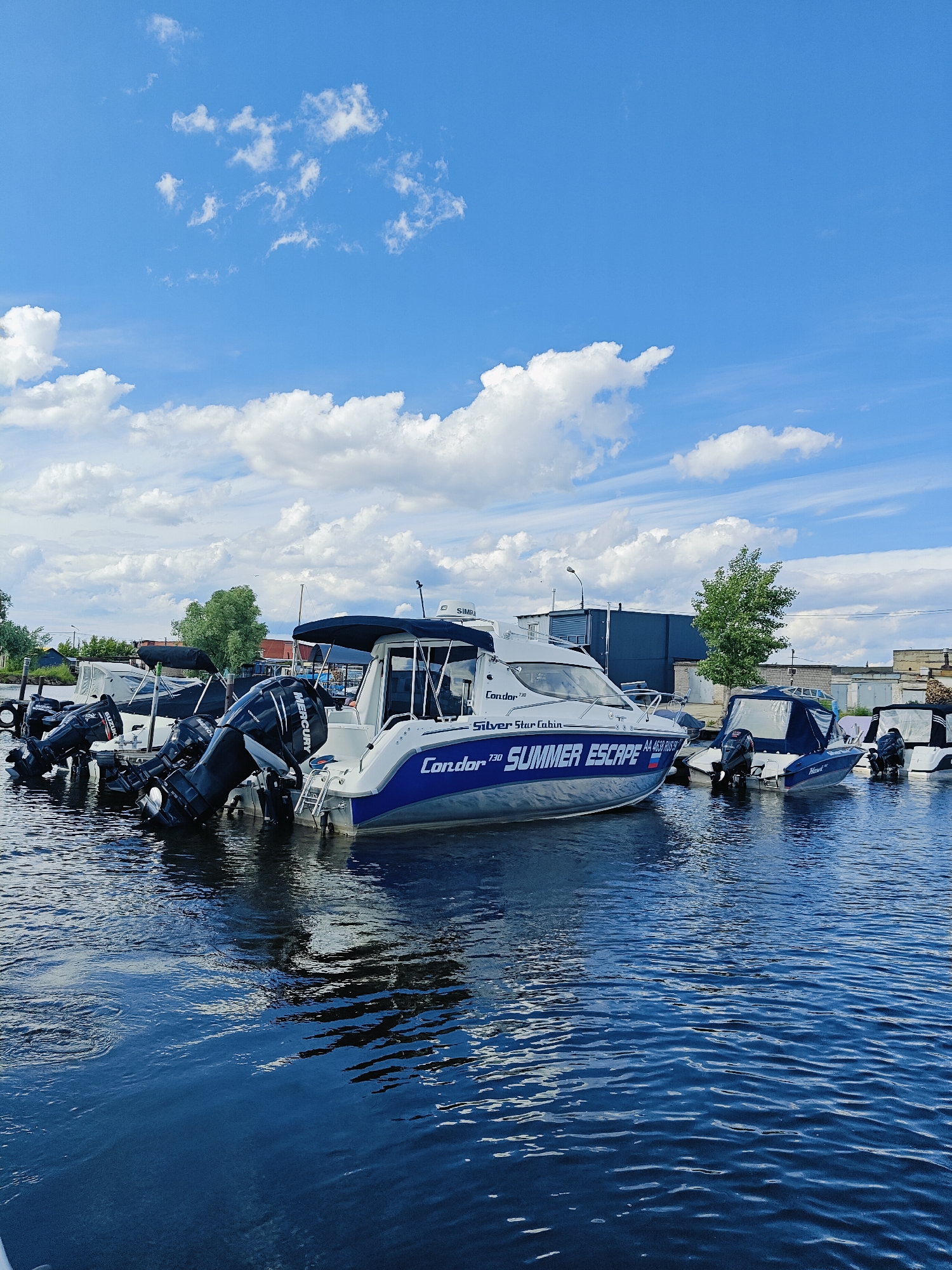 The image shows a body of water, likely a river or canal, in Chaikovsky, Russia. The water is dark blue with visible ripples. Several boats are docked or moored. The largest boat in the foreground is white with a blue stripe and the text "Condor 730" and "Summer Escape" visible on its side. It has two outboard motors. Other boats are visible behind it, some covered and some with outboard motors. The background features industrial buildings, trees, and a blue sky with scattered white clouds. The scene appears to be a marina or a boat storage area. There are no people visible in the image.