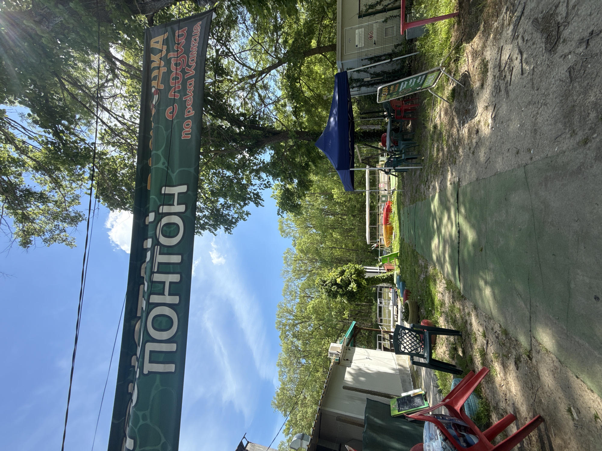 The image shows an outdoor area in Novo Oriahovo, Bulgaria. A green banner with white Cyrillic text is prominent, advertising "Ponton." The banner also contains the words "lodka s lodki po reka Kamzia." Adjacent to the banner is a dirt path leading towards trees. On the right of the path is a green artificial surface. Chairs and tables are scattered around, some under a blue canopy. There are buildings in the background. The sky is blue with light clouds.