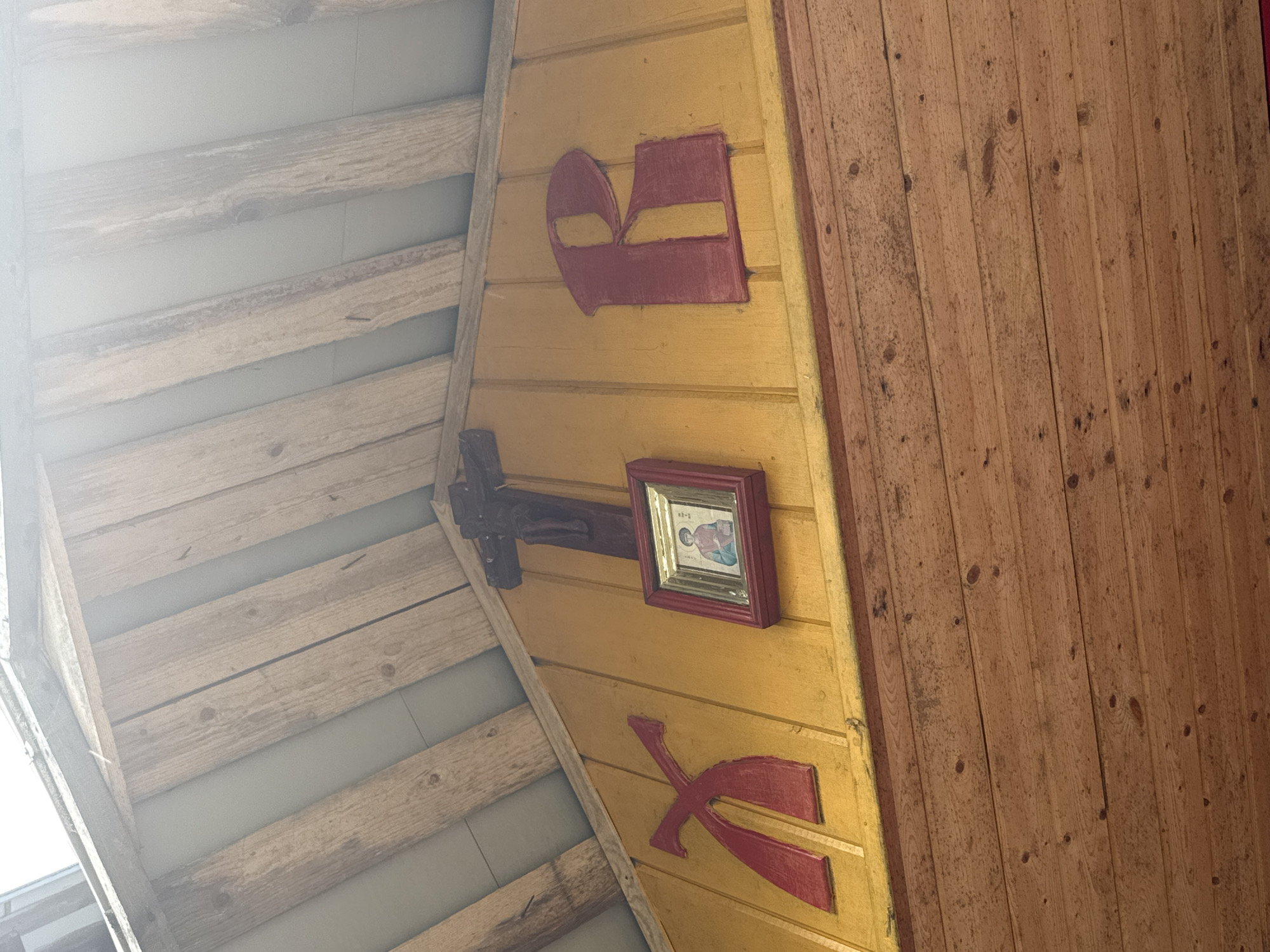 The image shows a close-up view of a yellow wooden wall with religious symbols. On the left side, there is a large red, stylized letter "X". To the right of it, a framed icon of a person is hanging, and above it, a wooden crucifix with a figure of Jesus. To the right of the icon, a large red letter "R" is mounted on the wall. The wooden wall is part of a building's exterior, with visible roof beams and wooden planks overhead. The lower portion of the image displays more wooden planks, likely part of the building's facade. The lighting suggests it is daytime, with sunlight filtering from the top. The overall scene suggests a religious or cultural structure in a rural or historical setting. This specific detail is located in Nenimyaki, Russia.
