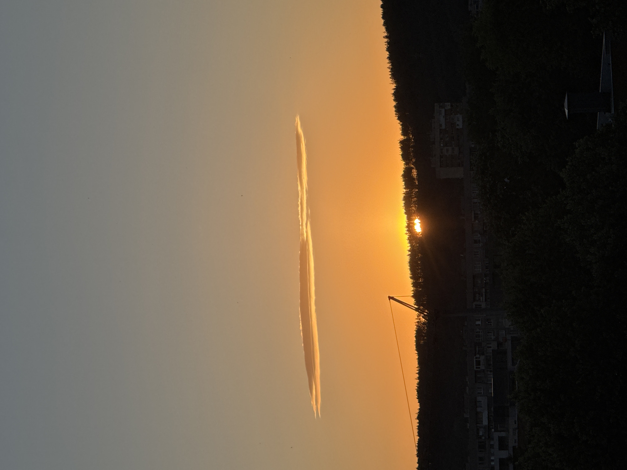 The image captures a sunset over the city of Pernik, Bulgaria. The sky displays a gradient of warm orange and yellow hues, transitioning to a lighter gray at the top. A distinctive, long, thin lenticular cloud is positioned horizontally in the upper portion of the sky, illuminated by the setting sun. The sun itself is partially visible, peeking through the silhouette of a tree-covered hill in the middle ground, casting a bright glow. A construction crane stands prominently on the left side of the image, silhouetted against the sunset. In the middle distance, apartment buildings are visible, some with multiple stories, also rendered in dark silhouette. The foreground is dominated by the dark shapes of trees and rooftops, with a chimney visible on a building in the lower right corner. The overall scene depicts an urban landscape at dusk.