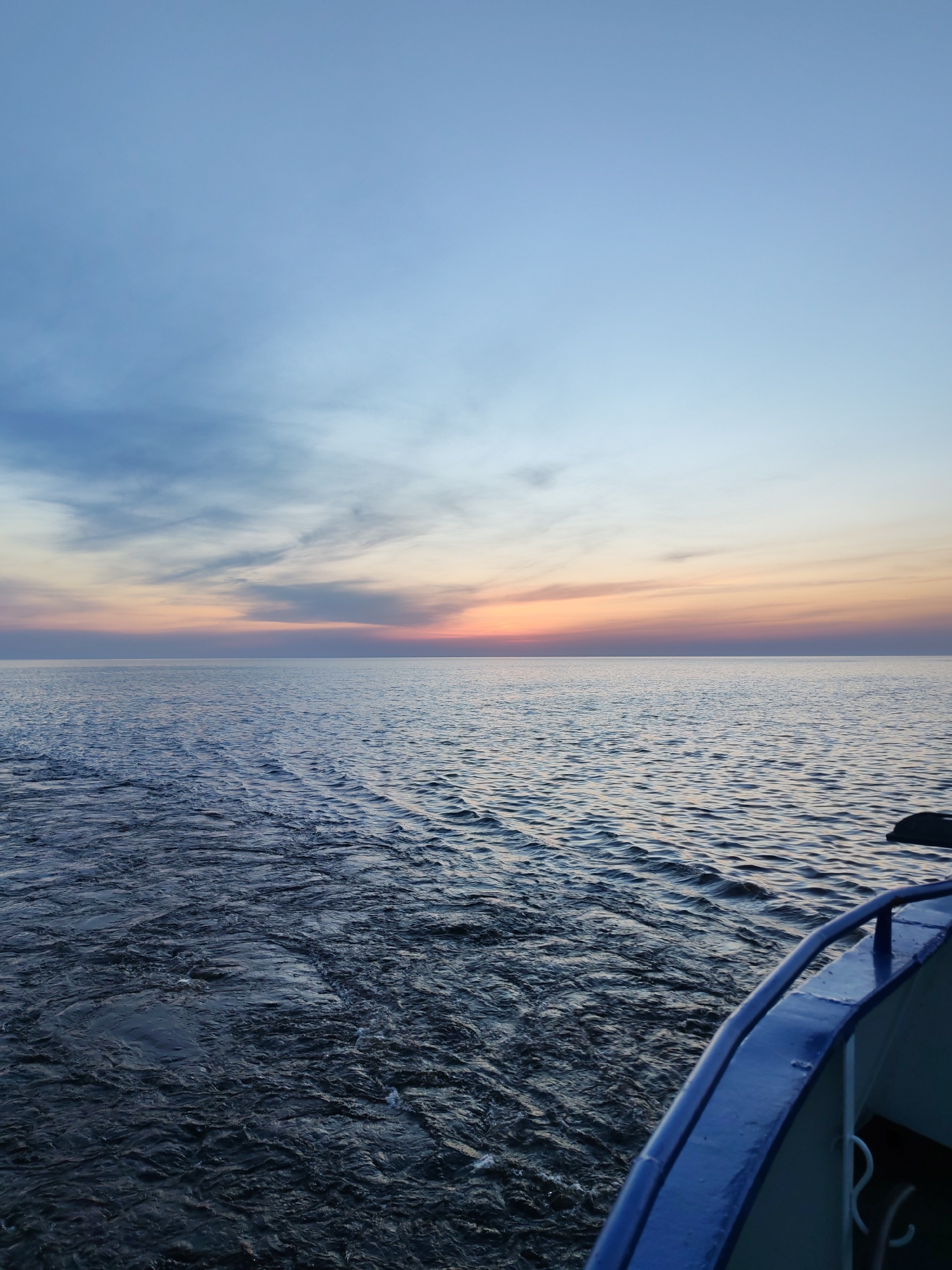 The image captures a seascape at sunset. The sky transitions from a soft blue to hues of orange and pink near the horizon. The sea is dark with ripples and waves, indicating movement. A portion of a blue boat railing is visible in the foreground, suggesting the photograph was taken from a vessel. The location is Unknown, Unknown.