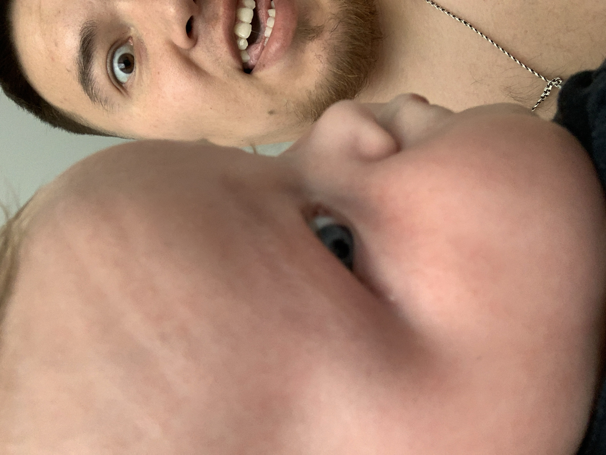 The image is a close-up, selfie-style shot featuring a baby in the foreground, whose face is partially out of focus and occupies the left side of the frame. The baby's skin is pink and smooth, with a hint of blonde hair visible at the top. To the right, a man's face is in sharp focus, positioned behind the baby. He has brown eyes, dark hair, and light stubble on his chin. His mouth is open, revealing teeth and tongue, with an exaggerated expression. He is wearing a silver chain around his neck. The background is a plain, light-colored wall. There is no identifiable information within the image to suggest a location in Stavropol, Russia.