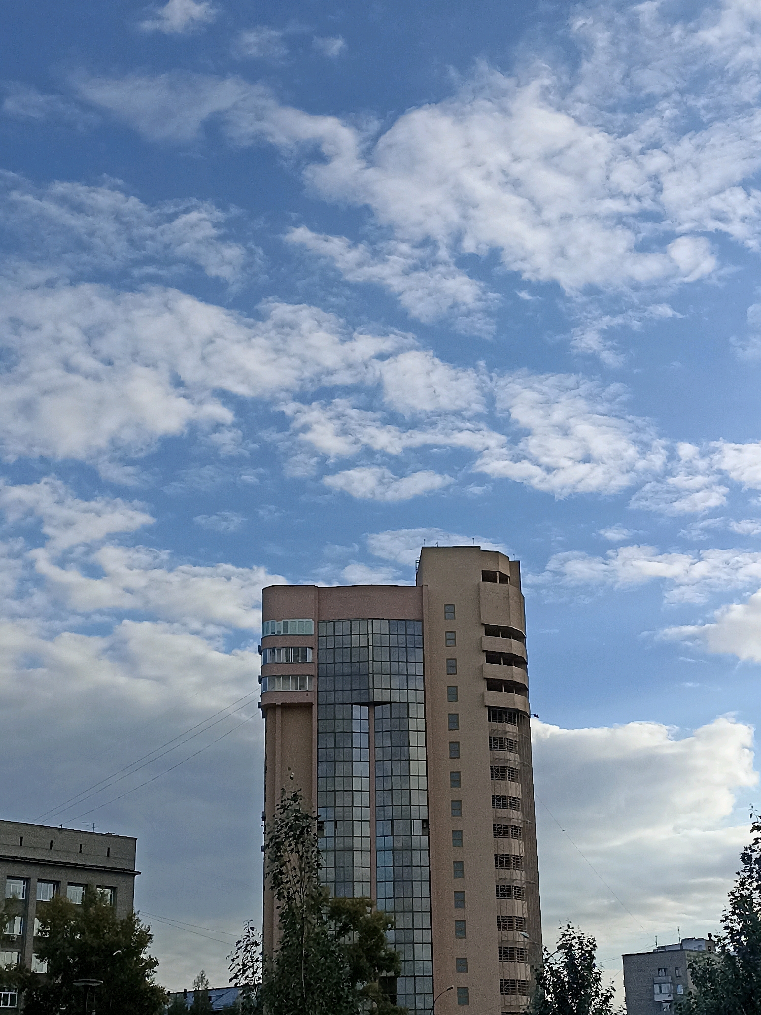 This is a vertical, eye-level, outdoor, daytime photograph of a tall, modern building under a blue sky with white clouds. The building is light brown and has a rounded section with multiple balconies. The main section of the building is glass-fronted, reflecting the sky and surrounding structures. To the left of the main building, a shorter, gray building with rectangular windows is visible. Trees with green foliage are in the foreground, partially obscuring the lower parts of the buildings. The scene appears to be in a city environment. This building is located at **Mikhailovskaya Street, 12, Novosibirsk, Russia.**