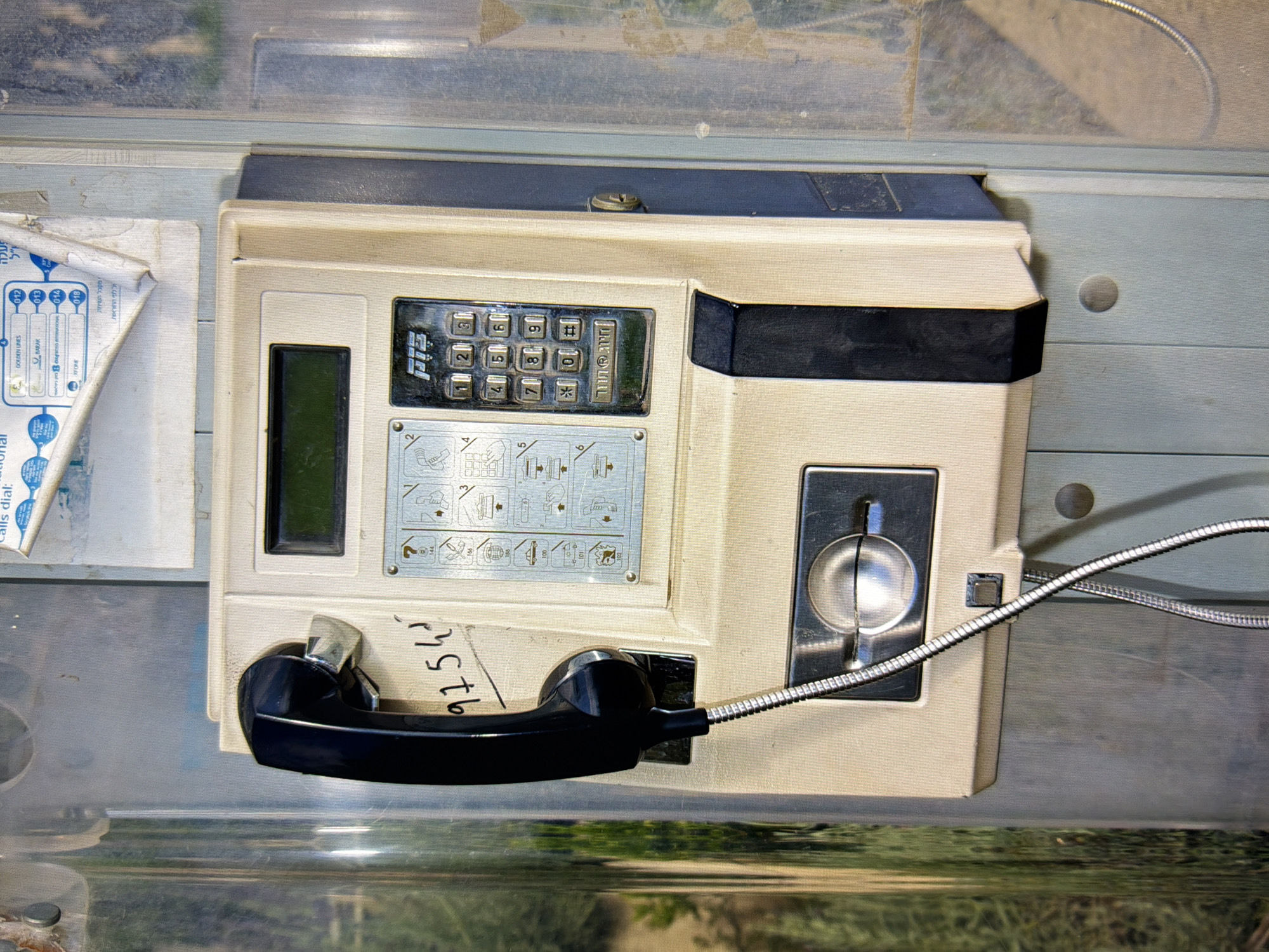 A close-up, eye-level shot captures a public payphone mounted on a grey, weathered pole, likely in Kyiv, Ukraine, given the context of similar images. The payphone unit itself is predominantly off-white and beige, with a prominent black handset resting on a cradle. A metallic coin slot is visible below the handset. The keypad features numbers 0-9, along with asterisk and hash symbols, and a logo that reads "PTS" in stylized silver lettering. To the left of the keypad, there's a small LCD screen. Instructional diagrams, showing how to use the phone, are adhered to the unit. A serial number, "9754", is handwritten on the unit in black ink. The background is blurred, showing hints of green foliage and indistinct urban elements, suggesting an outdoor setting. The overall condition of the payphone appears functional but aged, showing signs of wear and tear.
