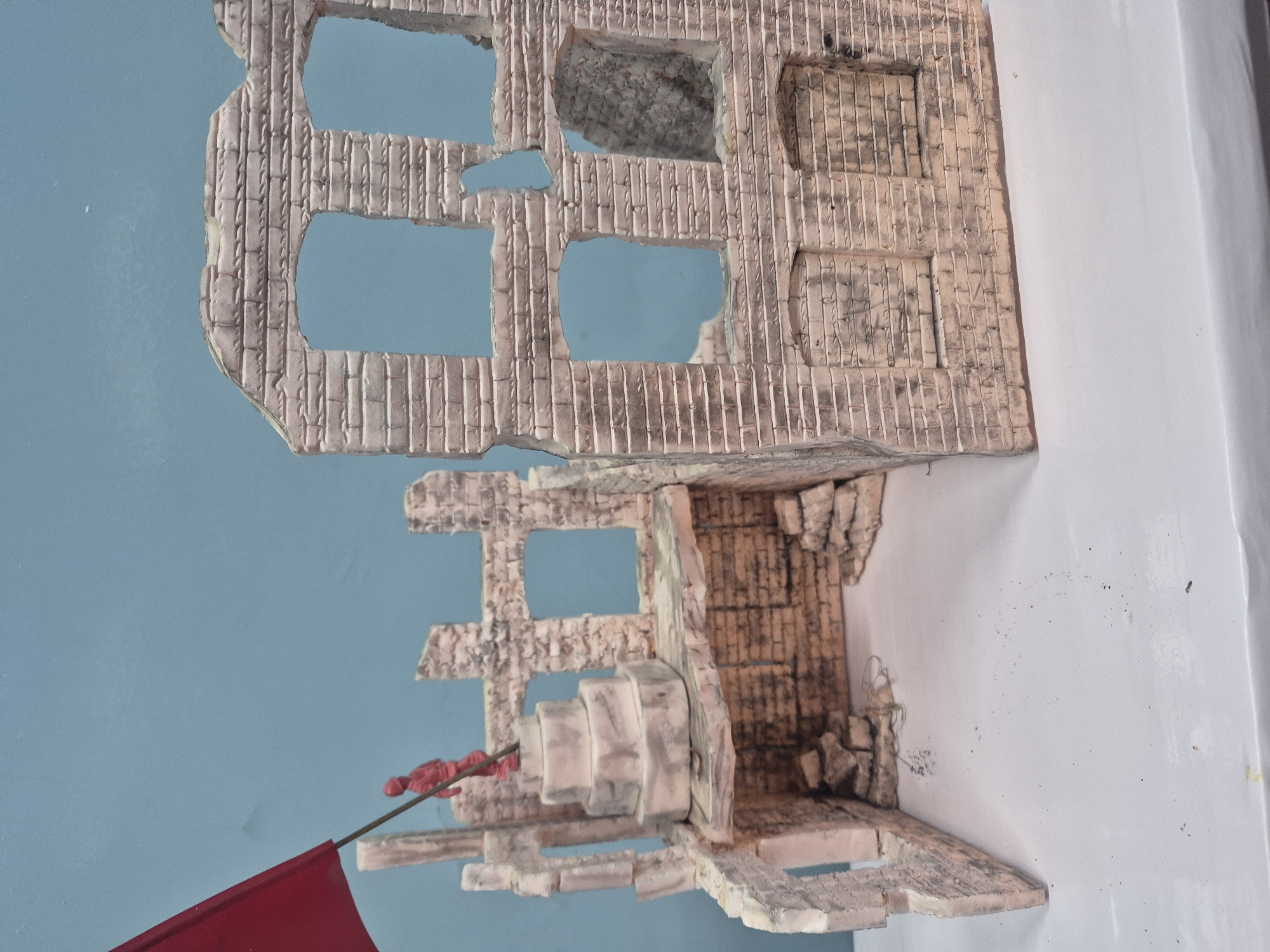 The image depicts a model of a war-torn brick building. The structure is incomplete, with gaping holes and broken sections of walls, revealing interior details. A red flag is planted on what appears to be a remnant of a staircase. The background is a two-toned blue and white, resembling a wall surface. The model's brickwork suggests the representation of a building in a conflict zone, potentially situated in a city, based on the context provided (Chaikovsky, Russia).