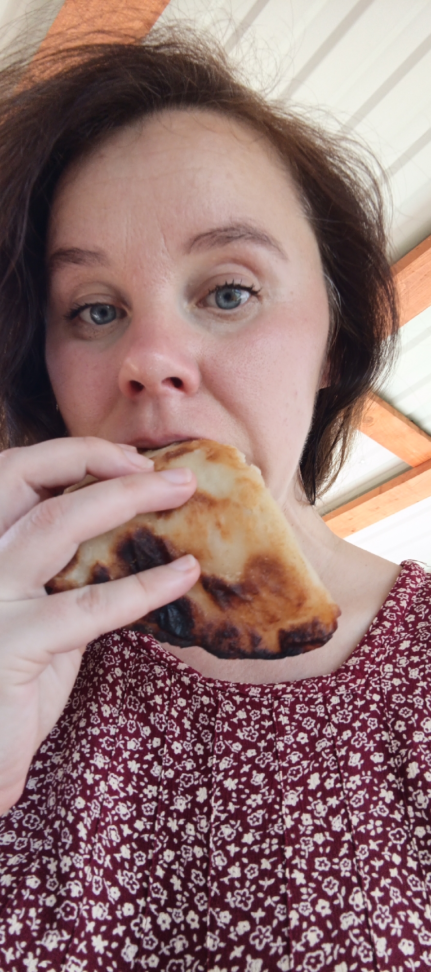 A medium close-up shot features an adult female looking directly at the camera while consuming a baked food item. Her hair is dark brown with visible wisps and some gray strands on the sides, and her eyes are blue. She has a fair complexion, with some redness around her nose and cheeks, and visible horizontal lines on her forehead. She is wearing a dark red garment with a repeating small white floral pattern, which appears to be pleated. With her left hand, she holds a golden-brown, irregularly shaped pastry or flatbread that has several distinctly charred, dark areas, particularly on its lower left portion. The food is partially obscured by her mouth, indicating the act of eating. The background consists of a light-colored, possibly corrugated metal or plastic ceiling or roof section, and light brown wooden structural beams visible on the right side, suggesting a sheltered outdoor or semi-outdoor environment. No information regarding a specific city or country location is discernible from the image itself.