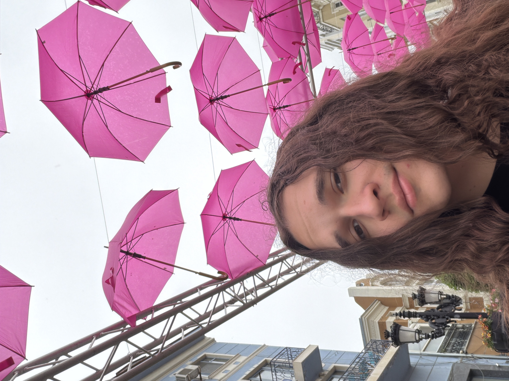 A low-angle selfie shows a person with brown, wavy hair and fair skin looking at the camera. Above the person's head are numerous pink umbrellas suspended in the air. The support structure for the umbrellas is visible. In the background, architectural details of buildings are discernible, including windows, decorative facades, and ornate street lamps. The location appears to be in an urban setting, likely within the city of Plovdiv, Bulgaria.