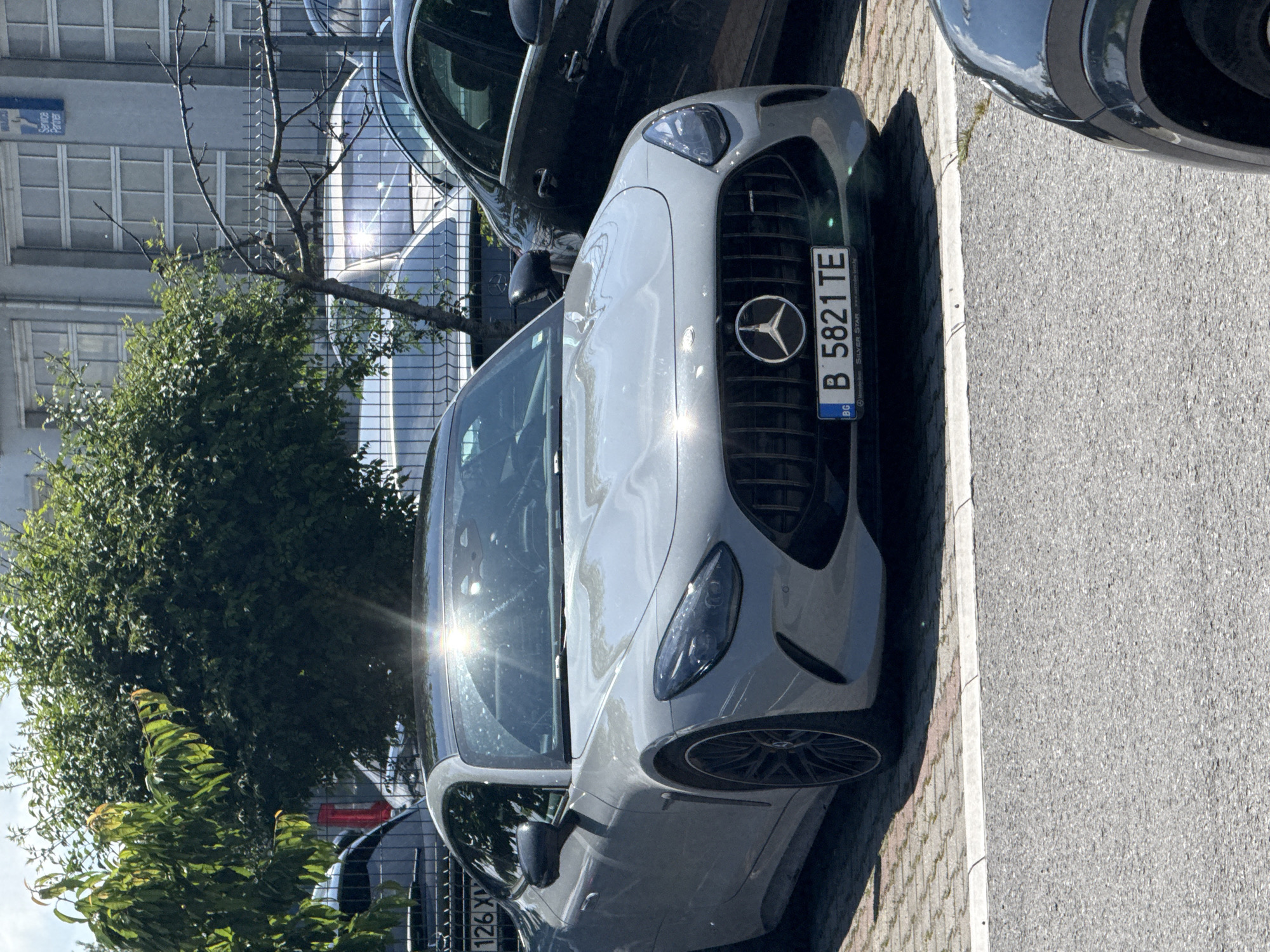 This image, captured during the daytime under sunny weather, depicts a street scene with several parked cars and lush greenery. The dominant subject is a light gray (possibly silver) Mercedes-Benz AMG GT, positioned prominently in the foreground. Its front fascia, featuring the iconic Mercedes-Benz star emblem on the grille and sleek headlights, is clearly visible. Bright sunlight creates strong reflections on the car's windshield and hood.
Behind the AMG GT, a darker-colored car, possibly an SUV, is partially visible. Further in the background, another dark vehicle is parked. On the left side of the image, behind the cars, green trees and bushes are flourishing, some bare branches are also visible. A modern building with multiple rectangular windows constitutes the background, reflecting the sunlight in some panes.
The vehicles are parked on a patterned paved surface, likely cobblestones or pavers, which is separated from an asphalt road by a light-colored curb. The scene suggests a typical urban or suburban environment.
Visible text includes the license plate on the Mercedes-Benz AMG GT, which reads "B 5821 TE" with "BG" (indicating Bulgaria) in a blue field on the left. The license plate frame also displays "Mercedes-Benz," "SILVER STAR," and the website "www.mercedes-benz.bg." A partially visible blue sign with white Cyrillic text, possibly "БАЛКАН," is affixed to the building in the background. While the context states Varna, the "B" on the license plate typically denotes Sofia, Bulgaria.