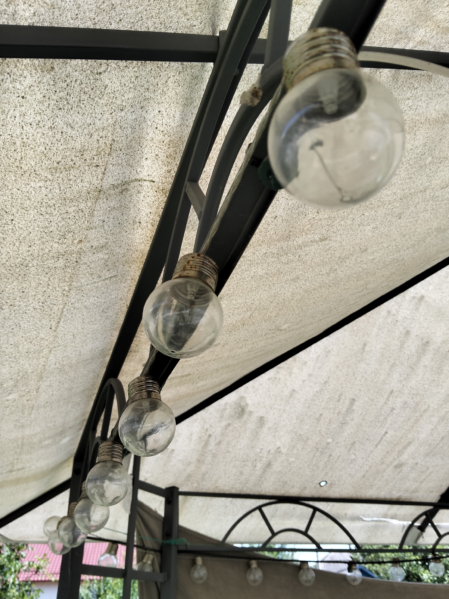 The image shows a string of outdoor globe lights hanging from a dark metal frame supporting a textured, off-white fabric canopy. The canopy appears aged and slightly dirty, with scattered dark specks. The lights are clear glass bulbs with visible filaments and corroded metal bases. They are strung in a downward-facing line, with the closest bulb in sharp focus and the others receding into a shallower depth of field. The background is blurred, but glimpses of greenery and a building with a red roof suggest an outdoor, possibly residential, setting in Ivancea, Moldova. The scene is captured from a low angle, looking up at the lights and canopy. There are no people visible in the image.