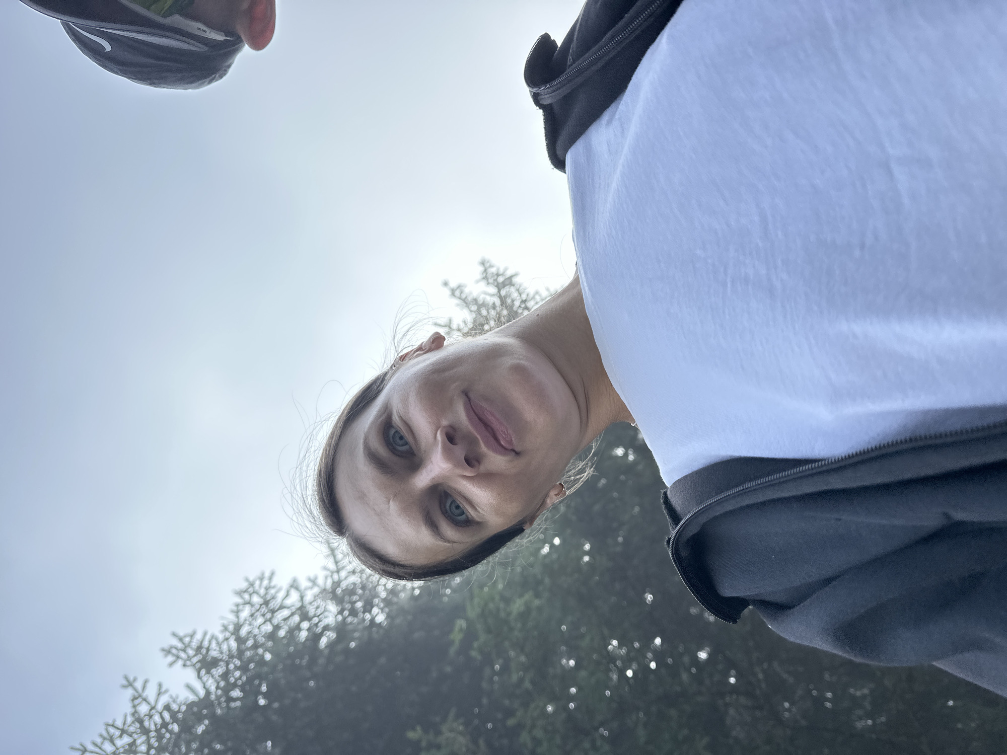 A low-angle, close-up, outdoor photograph captures a woman with light brown hair, her face turned towards the camera, eyes wide with a slight smirk. She is wearing a white t-shirt and a black zipped jacket. Behind her, blurred green foliage suggests a natural, possibly forested, environment. A portion of another person's head, wearing a black baseball cap, is visible in the upper right corner of the frame. The sky above is overcast and pale. This image appears to have been taken in Trešnjevik, Montenegro, given the context of the surrounding trees and the type of landscape typically found in that region.