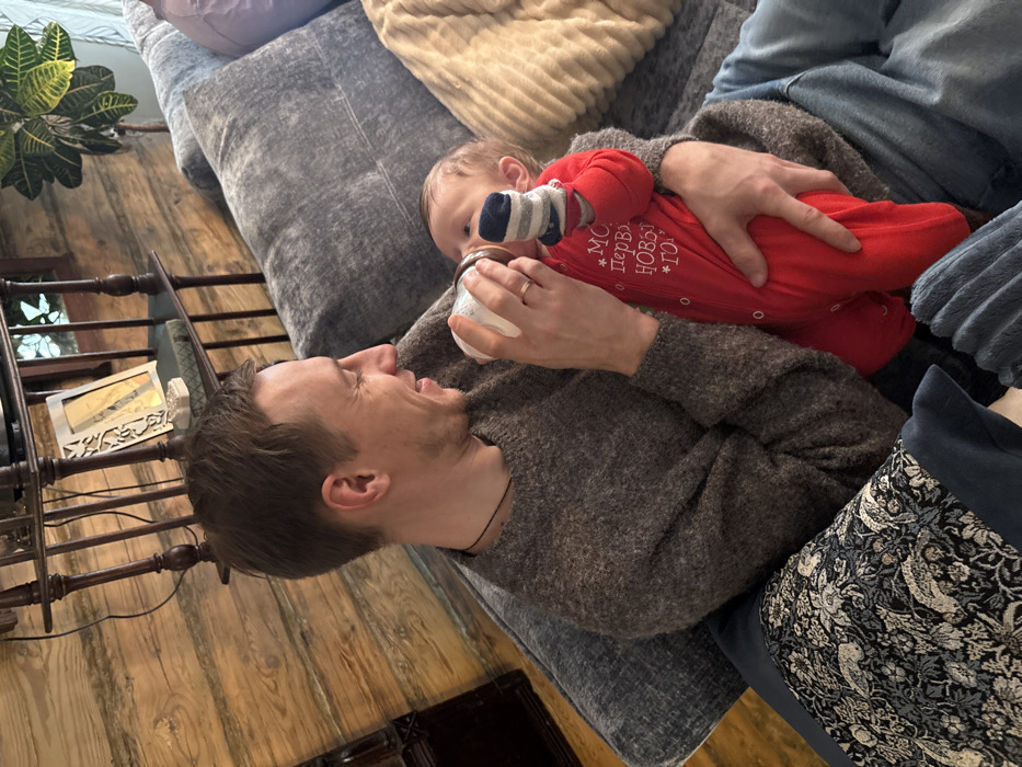 An adult male is seated on a grey fabric sofa, feeding an infant with a white baby bottle in a residence in Lezhnevo, Russia. The male, with short brown hair, wears a grey sweater and blue jeans, a thin dark necklace, and a ring on his left ring finger. He is smiling and looking at the infant. The infant wears a red romper with the Cyrillic text "МОЙ ПЕРВЫЙ НОВЫЙ ГОД" and striped socks, actively drinking from the bottle while being cradled by the male. The background features a log cabin-style wooden wall, a dark wooden shelf unit with decorative items, and a potted plant. The sofa has various throw pillows, including one with a dark blue bird and floral pattern and another light-colored, ribbed pillow.
