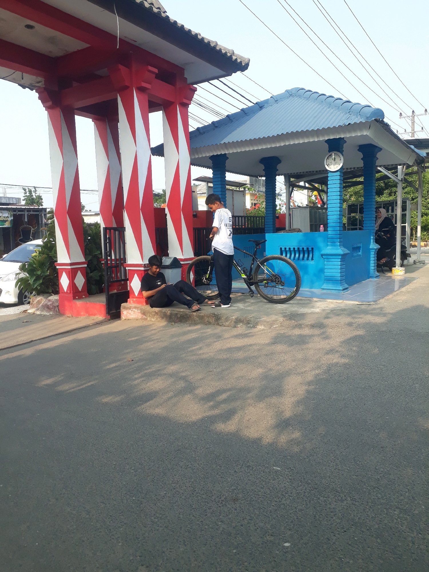 The image depicts an outdoor scene in the city of Pangkalpinang, Indonesia, possibly near a rest stop or public pavilion. In the foreground, a paved road is visible with tree shadows cast upon it. To the left, a red and white striped structure resembling an archway or gateway stands prominently. In front of this structure, a young man in a black t-shirt and pants is seated on a raised concrete step, looking at his phone. To his right, a bicycle with a green and black frame is parked.
Further into the scene, to the right of the red and white structure, is a bright blue covered pavilion with a corrugated roof. A clock with Roman numerals is mounted on one of the blue pillars supporting the pavilion. Two young people are standing near the bicycle. One, wearing a white t-shirt with text and dark pants, appears to be talking to the other who is partially obscured. In the background, a white car is parked near some bushes. To the far right, a woman wearing a headscarf and dark clothing is visible near the blue pavilion. The overall environment suggests a public space with pedestrian access and a recreational area.