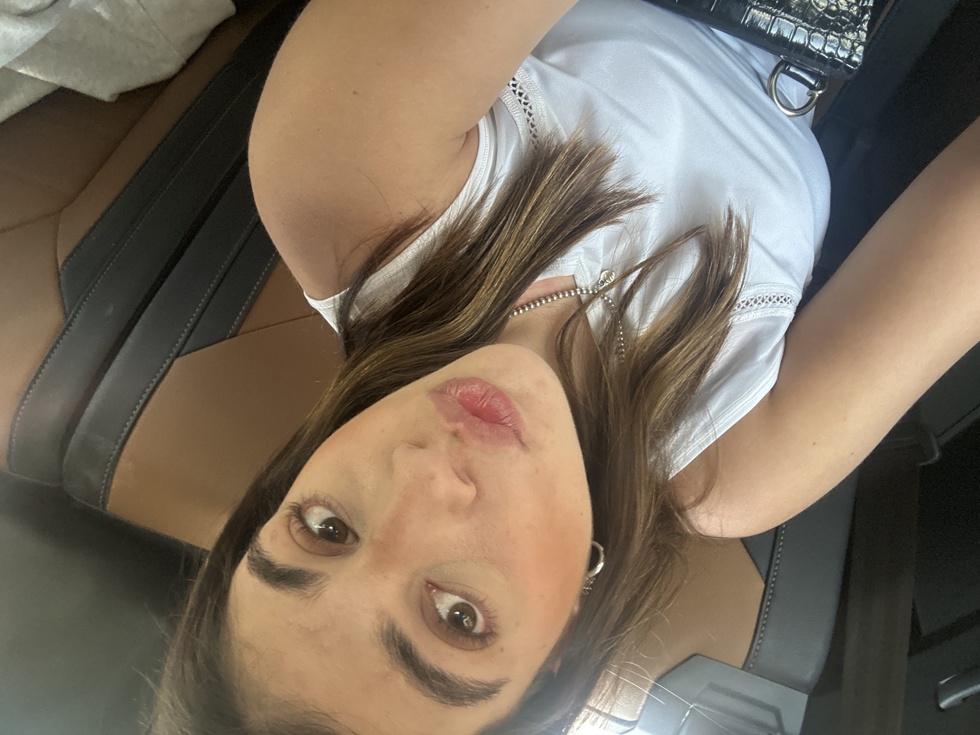 A close-up selfie-style image features a light-skinned female with brown hair. She is wearing a white tank top and a silver chain necklace. A black, textured bag is visible in the upper right corner of the frame. The background appears to be the interior of a car, with brown leather seats. Based solely on the image, it is not possible to pinpoint the specific location in Acapulco de Juárez, Mexico. There are no overt indications of activity, interaction, or environment that would allow for such localization within the city.