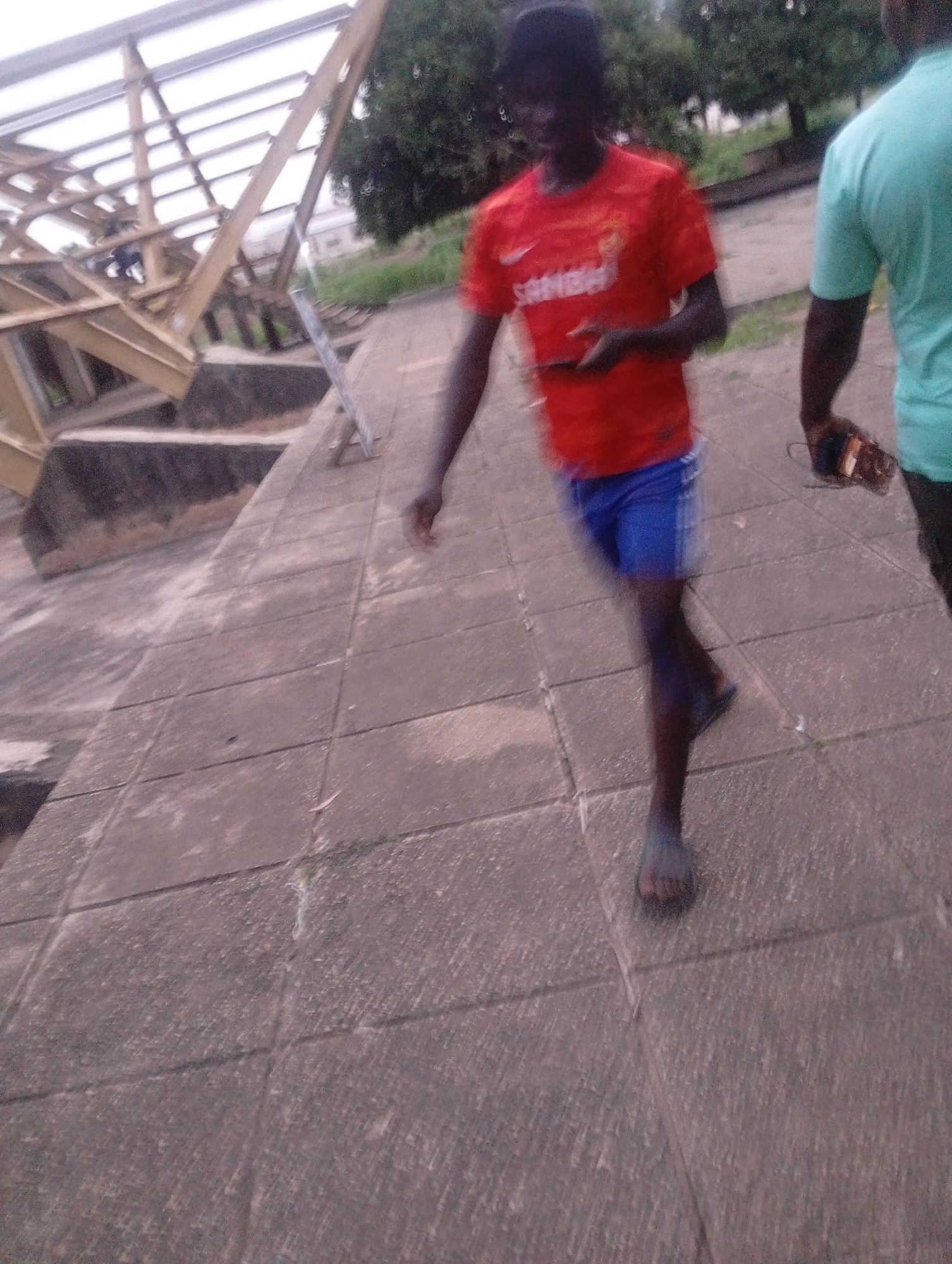 The image shows two young men walking on a paved walkway outdoors. The man in the foreground, wearing a red t-shirt with white text and blue shorts, is walking towards the right of the frame and looking down at his phone, which he is holding in his left hand. His right arm is extended slightly behind him. He is wearing sandals on his feet. Behind him and to his right, another man is walking, wearing a light blue-green t-shirt and dark pants. He is holding a brown object, possibly a wallet or small bag, in his right hand.
The setting appears to be an outdoor area with some trees and greenery in the background. A large, metallic structure with a corrugated roof is visible on the left side of the image, suggesting a covered walkway, stadium, or similar facility. The ground is paved with large, square concrete tiles, some of which show signs of wear and tear.
The lighting suggests it is daytime, possibly overcast, as there are no strong shadows. The overall mood seems casual, with the two individuals engaged in walking and presumably conversing or simply moving through the space. The image is slightly blurry, particularly of the foreground subject, indicating motion. The text on the red t-shirt is partially visible and appears to be "SAMBA" in white lettering. The location context is Jos, Nigeria.