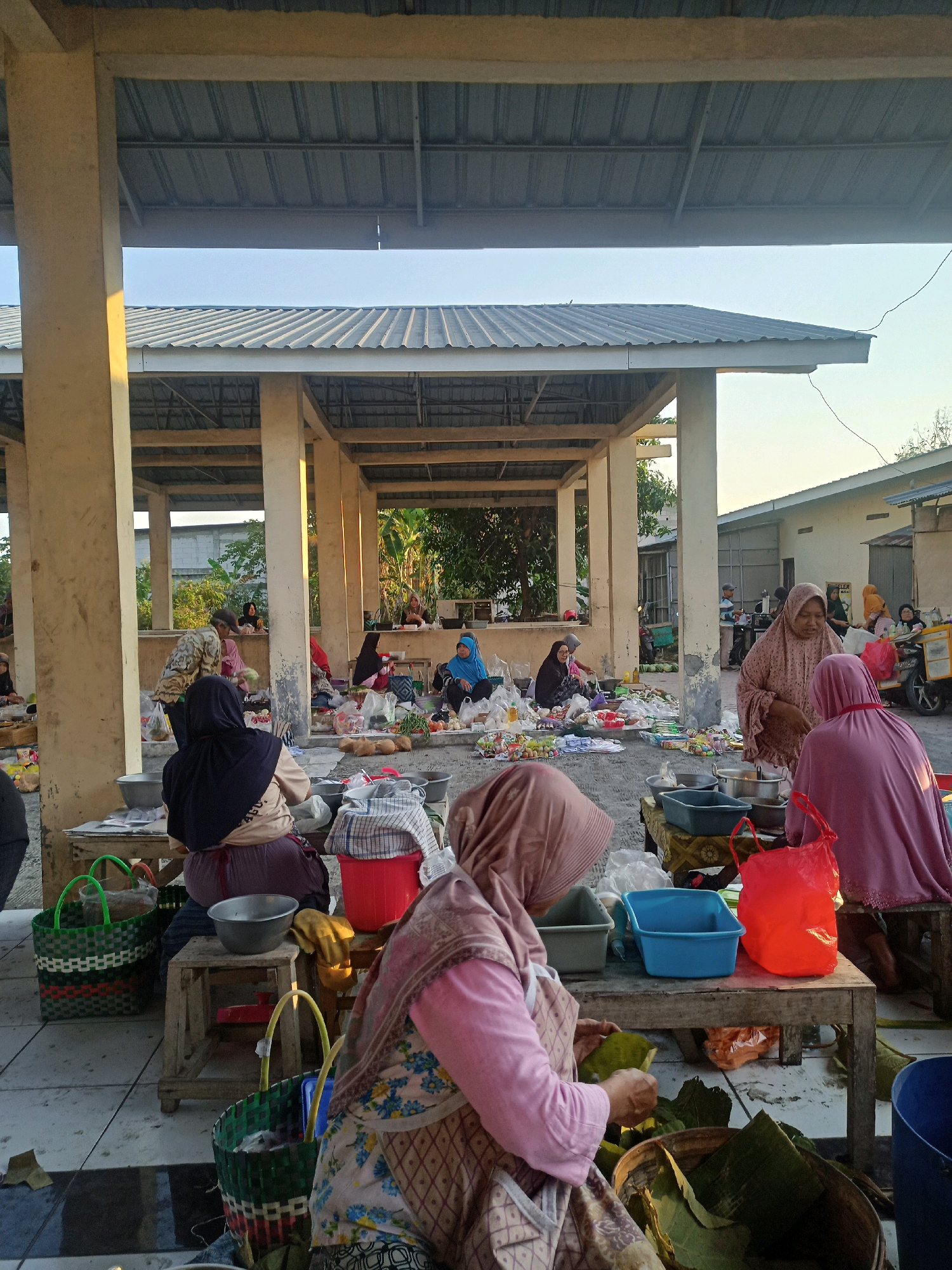 The image depicts a bustling outdoor market scene in Nganti, Indonesia, likely early in the morning or late afternoon given the warm, soft lighting. Several women, dressed in traditional Islamic attire with hijabs covering their hair, are actively engaged in selling and preparing goods. They are seated on the ground or low stools around makeshift stalls, which consist of wooden planks and tables. Various items are laid out for sale, including fresh produce like fruits and vegetables, and packaged goods. Some women are arranging their merchandise, while others appear to be serving customers or engaged in conversation. The market is sheltered by a utilitarian, open-sided structure with a corrugated metal roof supported by concrete pillars, a common sight in Indonesian markets. In the background, more market stalls and people can be seen, as well as some residential buildings and lush green foliage, suggesting the market is located in a semi-urban or rural setting. The overall atmosphere is one of lively commerce and community interaction.