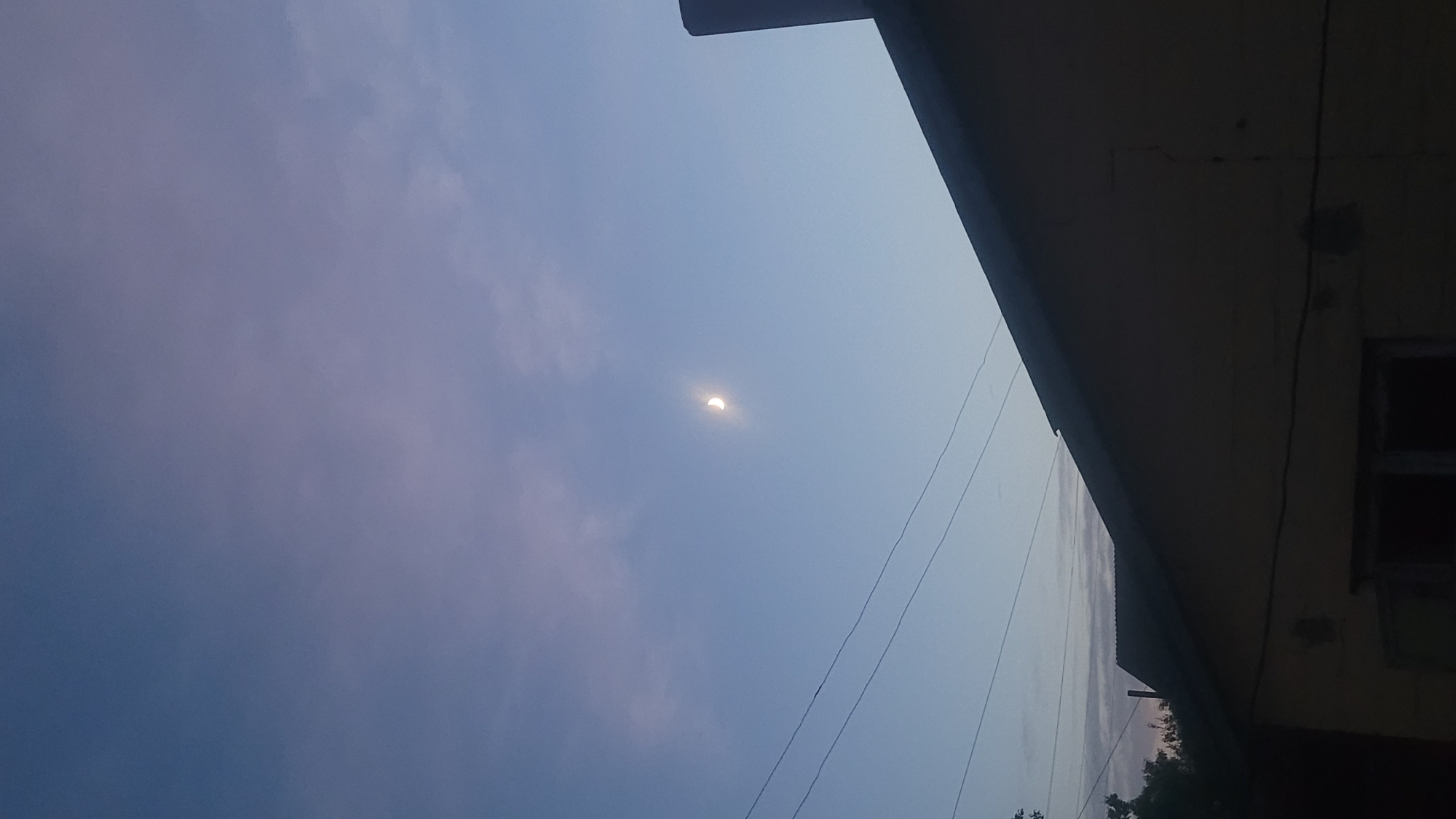 A crescent moon is visible in a pale blue and lavender sky. Three thin, dark wires stretch horizontally across the upper portion of the image. In the foreground, the dark roof of a yellow building slopes upwards from the left edge of the frame towards the right. Part of a dark, rectangular chimney or vent is visible on the far right, extending upwards from the roofline. The lower portion of the building is dark and indistinct, with what appears to be a window opening. Trees are visible on the far left edge of the frame, silhouetted against the sky. The overall impression is that of an outdoor scene at dusk or dawn.
