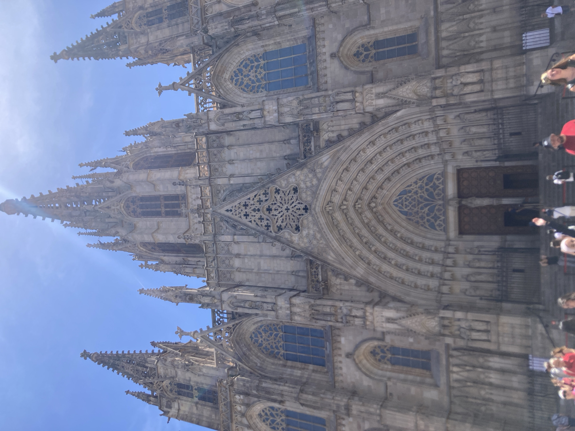 The image captures the grand facade of the Barcelona Cathedral, also known as the Cathedral of the Holy Cross and Saint Eulalia, located in the Gothic Quarter of Barcelona, Spain. The towering gothic architecture is dominated by intricate stone carvings, pointed arches, and numerous spires. Sunlight streams down from a clear blue sky, casting sharp shadows across the textured stone. In the foreground, a crowd of people are visible, appearing to be tourists visiting the landmark. Some are looking up at the cathedral, while others are engaged in conversation or walking. The overall scene depicts a sunny day at a prominent historical and religious site in Barcelona.