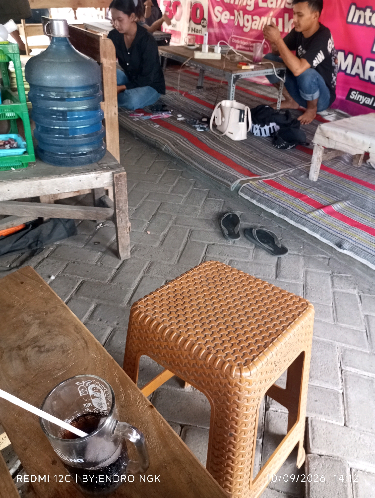 The image depicts a street scene in Nganjuk, Indonesia. In the foreground, a clear glass mug filled with a dark beverage and a white straw rests on a wooden surface. In the middle ground, a woven plastic stool is visible. Behind the stool, a blue water dispenser is situated next to a wooden table. Two individuals are present: one, a woman wearing a black top and blue jeans, is seated on the ground. The other, a man in a black t-shirt and blue jeans, is also crouched down, holding a phone. Various objects are scattered around, including flip-flops, a white bag, and a folded piece of cloth. The ground is paved with grey interlocking bricks, and a striped mat is laid out. In the background, a pink banner with text is visible. The overall setting suggests a casual, outdoor environment.