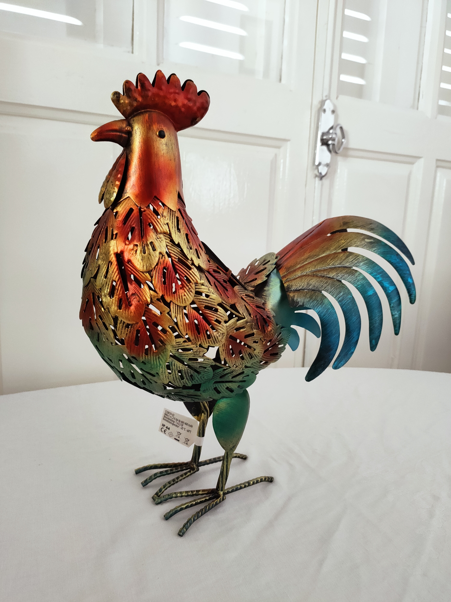 A decorative metal rooster, primarily red, gold, and green, stands on a white tablecloth. The rooster has a prominent red comb, a metallic beak, and a body adorned with intricate, feather-like cutouts. Its tail feathers display a gradient of colors, transitioning from brown to blue. The rooster is depicted in a side profile, facing left. In the background, a white door with horizontal paneling and a silver handle is visible. The scene appears to be indoors, possibly in a home or a shop, rather than a specific outdoor location in Ariana, Tunisia. No people are present in the image.