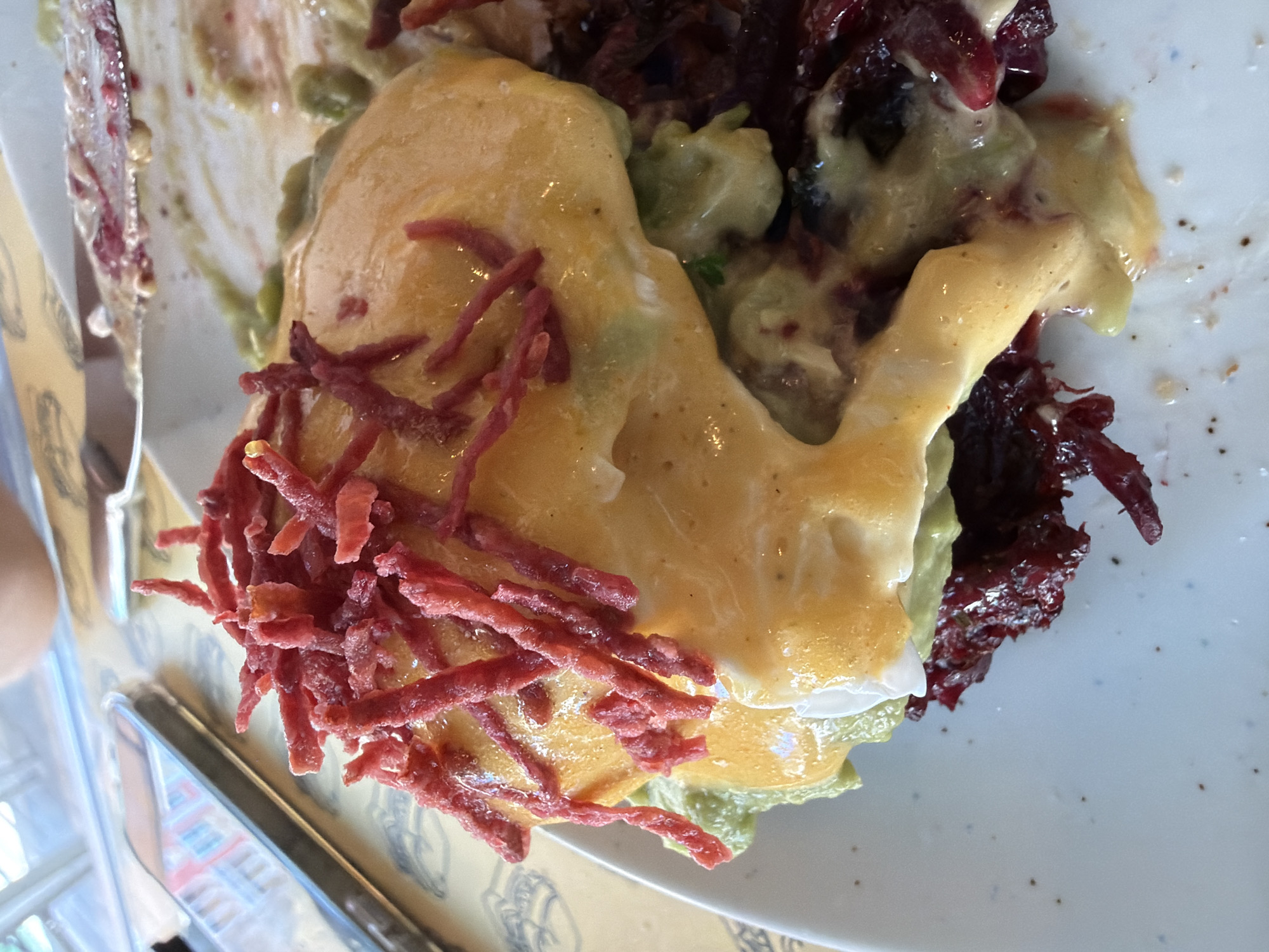 A close-up, overhead shot of a plate of food on a table, likely in a restaurant. The dish appears to be a modern take on eggs Benedict, featuring poached eggs covered in a rich hollandaise sauce, served over shredded beets and possibly avocado. Thin, red, shredded pieces, possibly a type of cured meat or vegetable, are sprinkled on top of the hollandaise. A fork and knife, with remnants of the food on them, are visible at the top of the frame, suggesting the meal is either being eaten or has just been served. The table surface beneath the plate has a yellow and white patterned tablecloth, and a smartphone is partially visible on the left side of the image. The lighting suggests an indoor setting, possibly a brightly lit cafe or restaurant. The composition is focused tightly on the food, with the background slightly blurred. There is no indication of the specific location within Doha, Qatar.