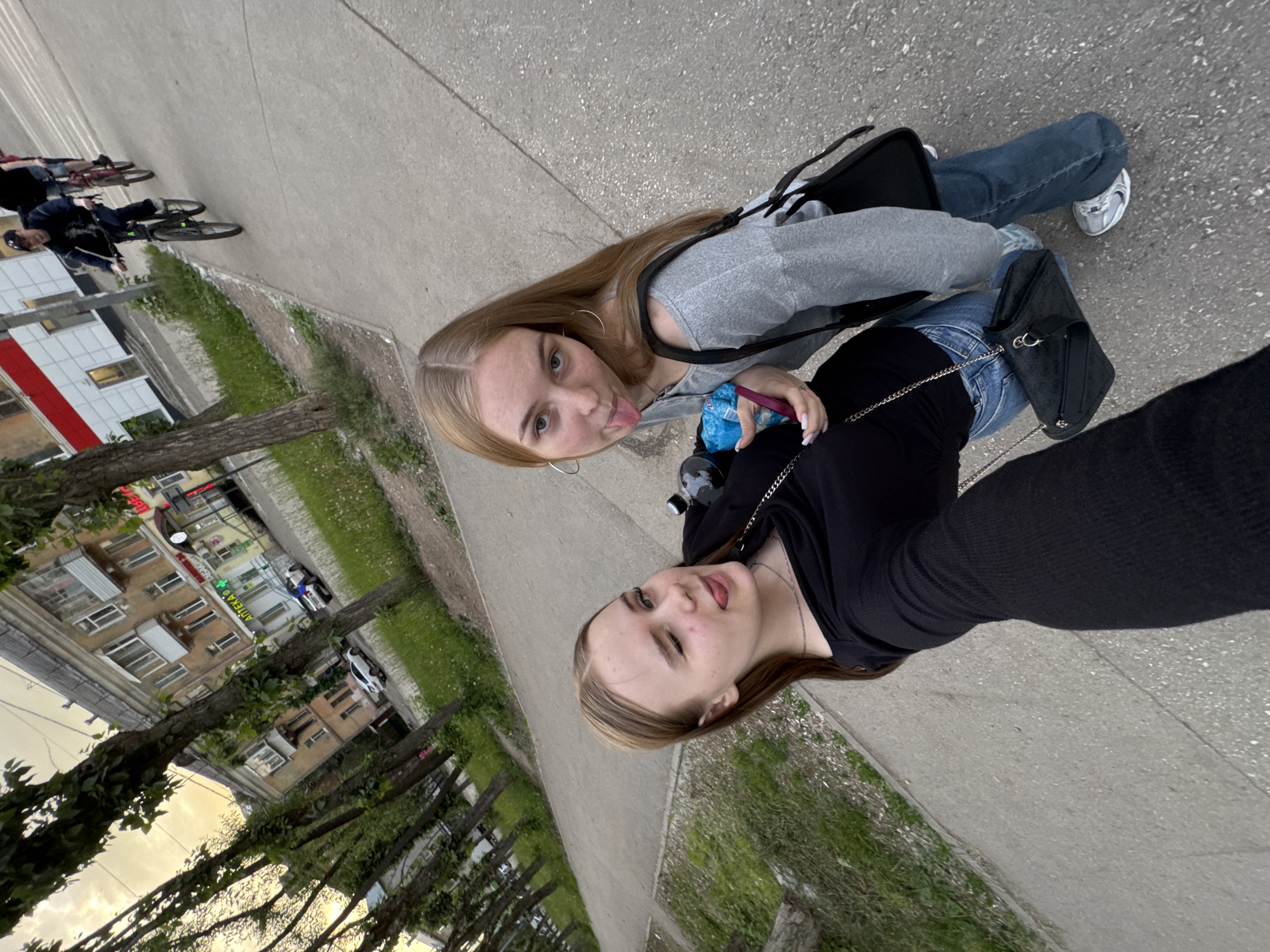 The image presents a slightly tilted, eye-level perspective of two young females posing for a selfie on an urban street in Perm, Russia, with background elements characteristic of a city environment.
In the foreground on the left, a female with light brown hair is seen winking with her right eye and sticking out her tongue. She wears a black long-sleeved, ribbed top, a silver chain necklace, and blue jeans, with a small black shoulder bag featuring a silver chain strap visible over her hip. She holds a black bottle in her left hand, partially obscured.
To her right, a second female with long, straight blonde hair and large silver hoop earrings also sticks out her tongue and looks towards the camera. She wears a grey off-the-shoulder long-sleeved top, blue jeans, and white sneakers with grey accents. She carries a black shoulder bag with a wider strap and holds a blue plastic package or bag, along with a small pink object, in her hands.
The setting is an urban street. A concrete sidewalk occupies the immediate foreground and middle ground, with an asphalt road running parallel to it in the background. On the road, three males are observed riding bicycles away from the camera. The lead cyclist wears a dark long-sleeved shirt, dark pants, and a dark baseball cap, riding a dark mountain bike.
Along the far side of the road, multi-story buildings are visible. One building exhibits a red and white facade on its lower section. Adjacent to it, an older-style building features several balconies and displays signage in Russian, including "АПТЕКА" (Pharmacy) in green lettering and "ЦВЕТЫ" (Flowers) in red lettering. Two parked cars, a white sedan and a dark sedan, are situated in front of these buildings. A row of mature deciduous trees with green foliage lines the sidewalk, and patches of grass interspersed with white dandelions are present between the trees and the buildings. The sky appears overcast with a soft, warm light, indicating either late afternoon or early evening.