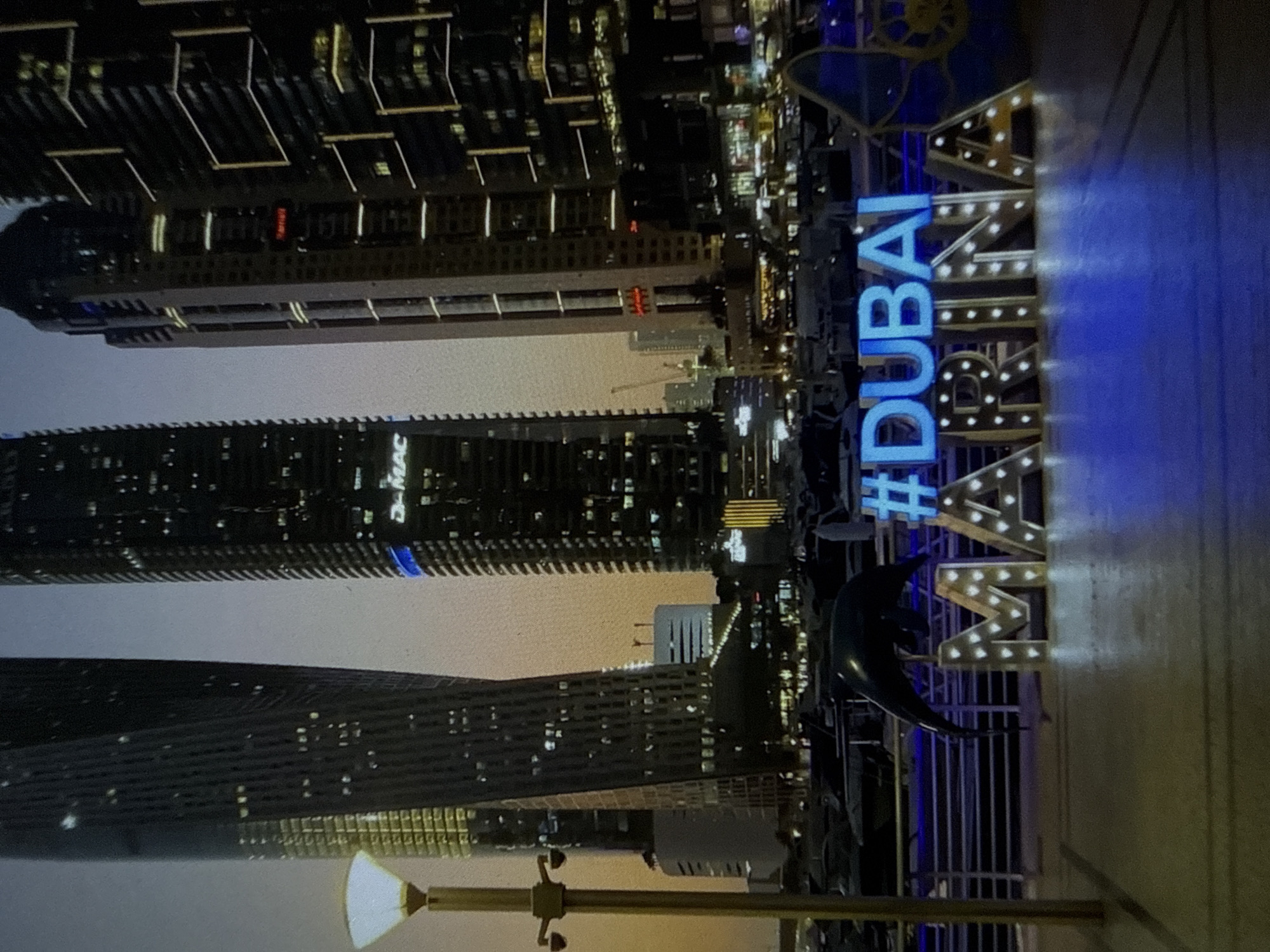 The image depicts a nighttime cityscape, appearing to be taken in portrait orientation and rotated 90 degrees clockwise. Towering, illuminated skyscrapers dominate the background, featuring numerous lit windows and architectural lighting. The sky is dark, indicating night.
In the foreground, on the right side of the image, a large, prominent illuminated sign spells out "#DUBAI MARINA". The "#DUBAI" portion is rendered in bright blue, possibly neon or LED lighting, while "MARINA" is spelled with individual, white, bulb-lit letters. To the left of the "MARINA" sign, a dark, crescent moon-shaped sculpture or decorative element is visible. A lamppost with a glowing light source is present in the lower left foreground.
The buildings display various architectural styles, with some having intricate facade lighting. On one of the buildings, faint white lettering that appears to read "D-MAC" is visible, possibly a company name or part of a building's title. Another building shows a red illuminated sign, but the text is not clearly discernible.
The scene conveys the vibrant, modern atmosphere of a major urban center at night. The distinct "#DUBAI MARINA" sign serves as a strong location cue, indicating the setting is Dubai Marina. This contradicts the provided location context of Varna, Bulgaria.