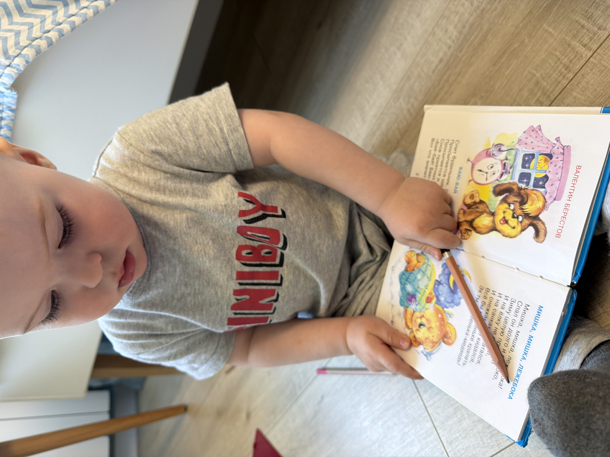 A child is sitting on the floor, holding open a book titled "ВАЛЕНТИН БЕРЕСТОВ" and "МИШКА, МИШКА, ЛЕЖЕБОКА". The child is wearing a gray t-shirt with the word "INIBOY" on it and is holding a pencil. The book is open to a page with illustrations of cartoon animals. The location appears to be indoors, possibly a home. The text within the book is in Russian. It is unknown if this is Votkinsk, Russia.