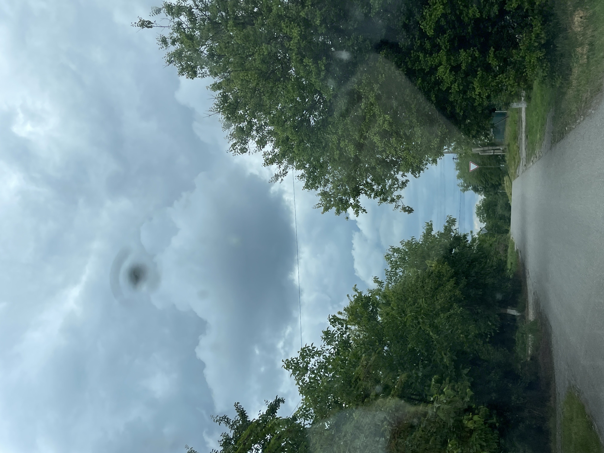 The image captures a rural road in Levokumka, Russia, under a cloudy sky. On the left side of the road, a dense line of green trees lines the path, with some branches extending over the road. On the right, another line of trees creates a similar natural boundary, and further down, a white triangular road sign is visible. The road itself appears to be asphalt, with a slight sheen indicating it might be wet. The sky is dominated by thick, gray clouds, with a peculiar, dark, circular anomaly visible in the upper center of the frame, resembling a "hole punch" cloud formation or a lenticular cloud. There are no people or animals visible in the image, and no actions are being performed. The overall impression is one of a quiet, overcast day in a rural setting.