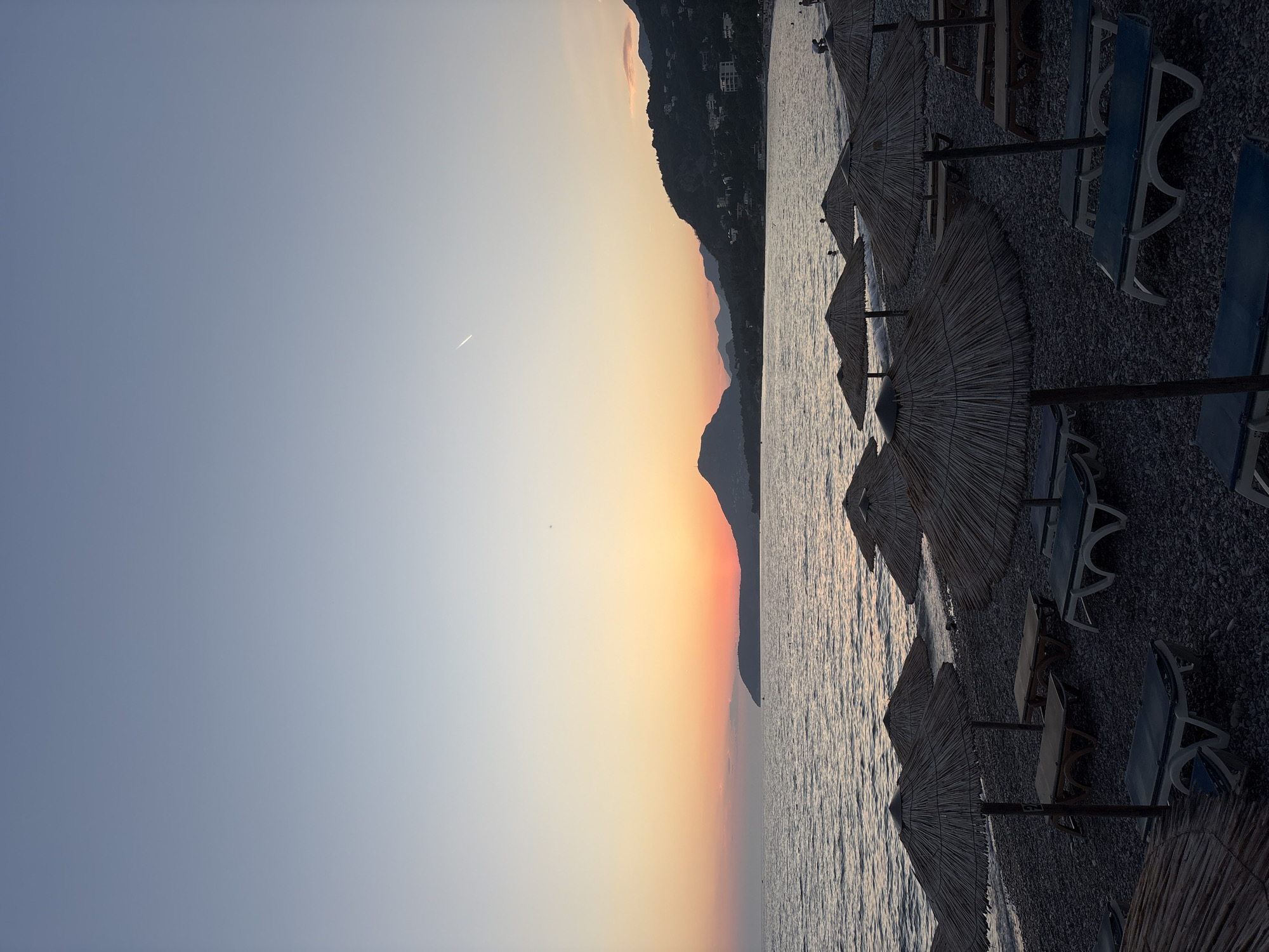The image depicts a serene beach scene in Bar, Montenegro, during sunset. The sky transitions from a clear, pale blue at the top to a warm gradient of orange and pink near the horizon, where a mountain silhouette is visible. The sea is calm, with gentle waves lapping at the pebble-strewn shore. Rows of thatched umbrellas and empty lounge chairs are arranged on the beach, suggesting a quiet end to the day. In the distance, the coastline is dotted with buildings, and the slopes of the mountains are covered in lush greenery. A faint contrail from an airplane stretches across the sky. A few people are sparsely scattered on the beach and in the water, enjoying the fading light.
