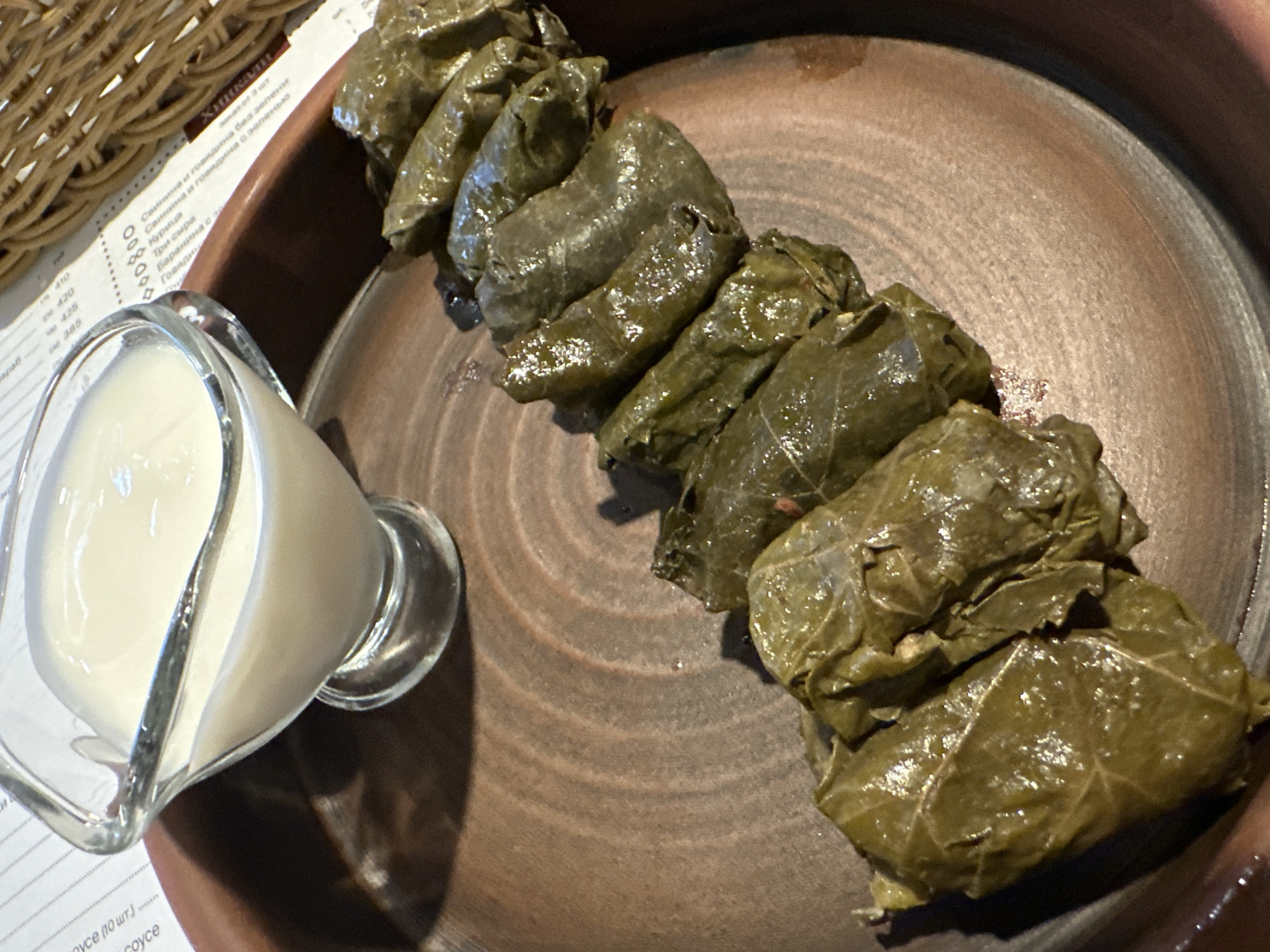 A close-up, high-angle shot captures a traditional meal presented on a rustic table setting. The primary subjects are a row of dolma (stuffed grape leaves) and a small glass pitcher of white, creamy sauce, likely sour cream or yogurt.
The dolma, dark green and glistening with moisture, are neatly arranged on a round, brown, unglazed ceramic plate that features concentric circular patterns. The plate's earthy tone provides a natural backdrop for the dish. To the left of the plate, the glass pitcher filled with the white sauce sits, its transparent material allowing the creamy texture to be visible.
In the background, a portion of a printed menu in Russian is visible, partially obscured by a woven basket. The menu lists various food items, most notably "Хинкали" (Khinkali), a type of Georgian dumpling, with different fillings such as "Свинина и говядина без зелени" (Pork and beef without greens), "Свинина и говядина с зеленью" (Pork and beef with greens), "Курица" (Chicken), and "Три сыра" (Three cheeses). Other partially visible text includes "Coyce" (Sauce) and numbers like 410, 420, 425, 240, 385, which likely indicate prices. The presence of Khinkali and dolma, along with Russian text, suggests a restaurant serving Caucasian or Eastern European cuisine, fitting the provided context of Saratov, Russia. The overall scene conveys a prepared meal, ready to be enjoyed.